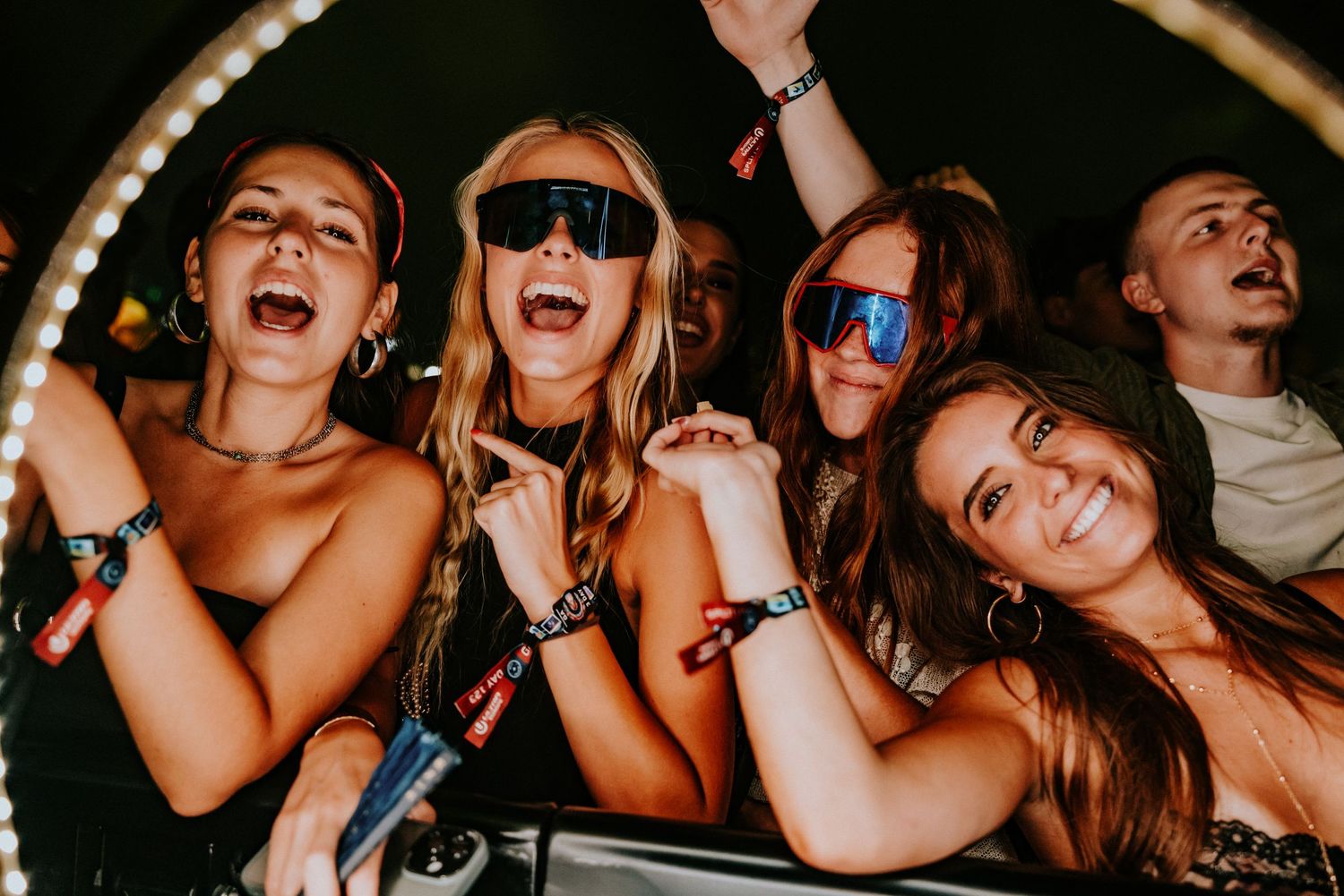 Excited young women wearing sunglasses cheering at an outdoor music festival concert at night.