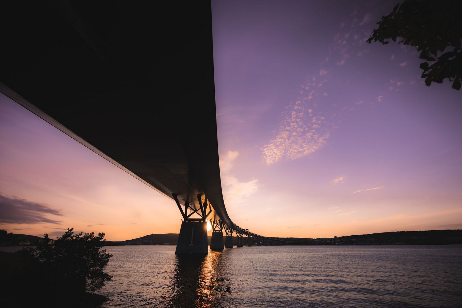 Panoramic sunset view over water with bridge silhouette and purple sky gradient.