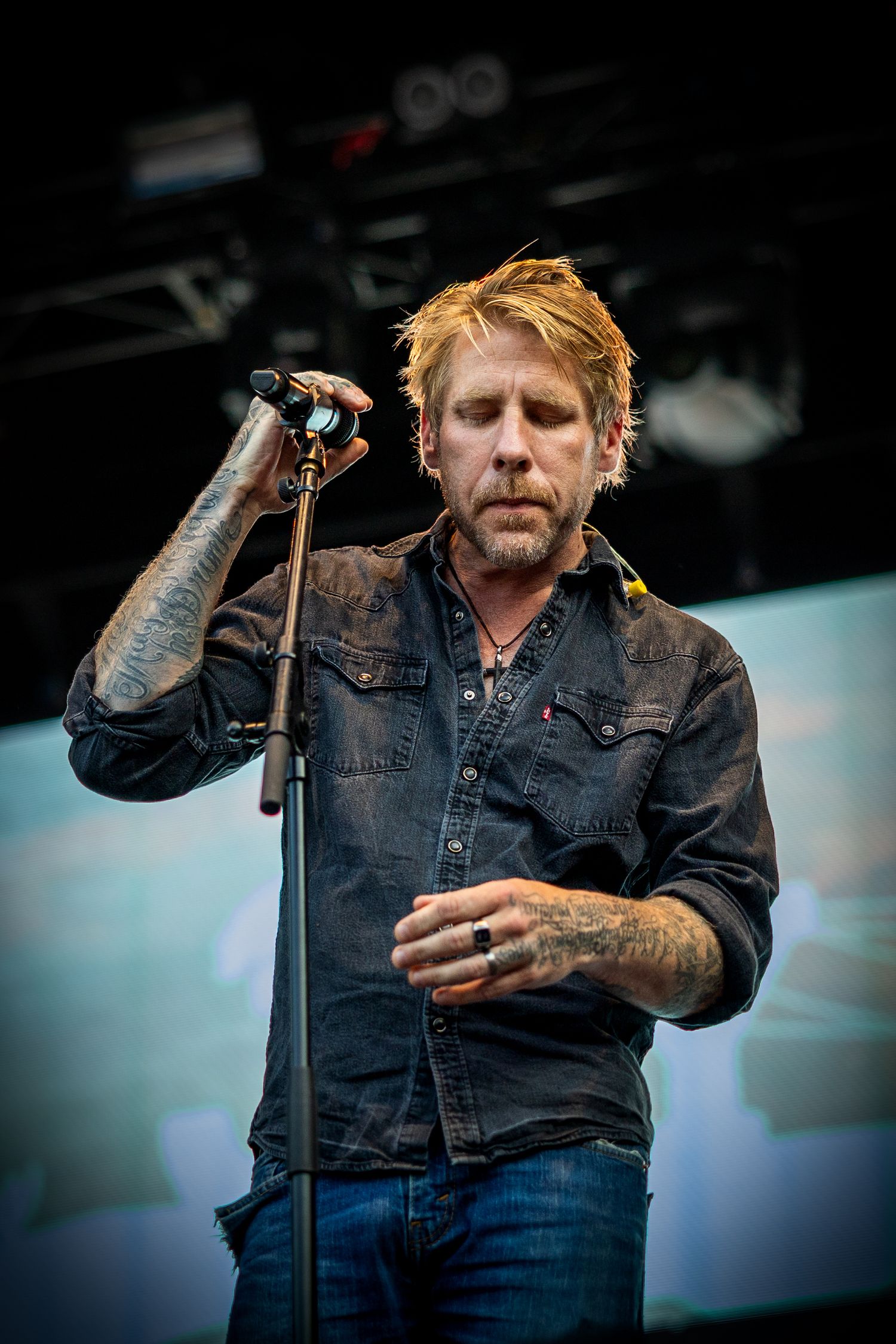 Performer in denim jacket on stage during daytime festival performance.