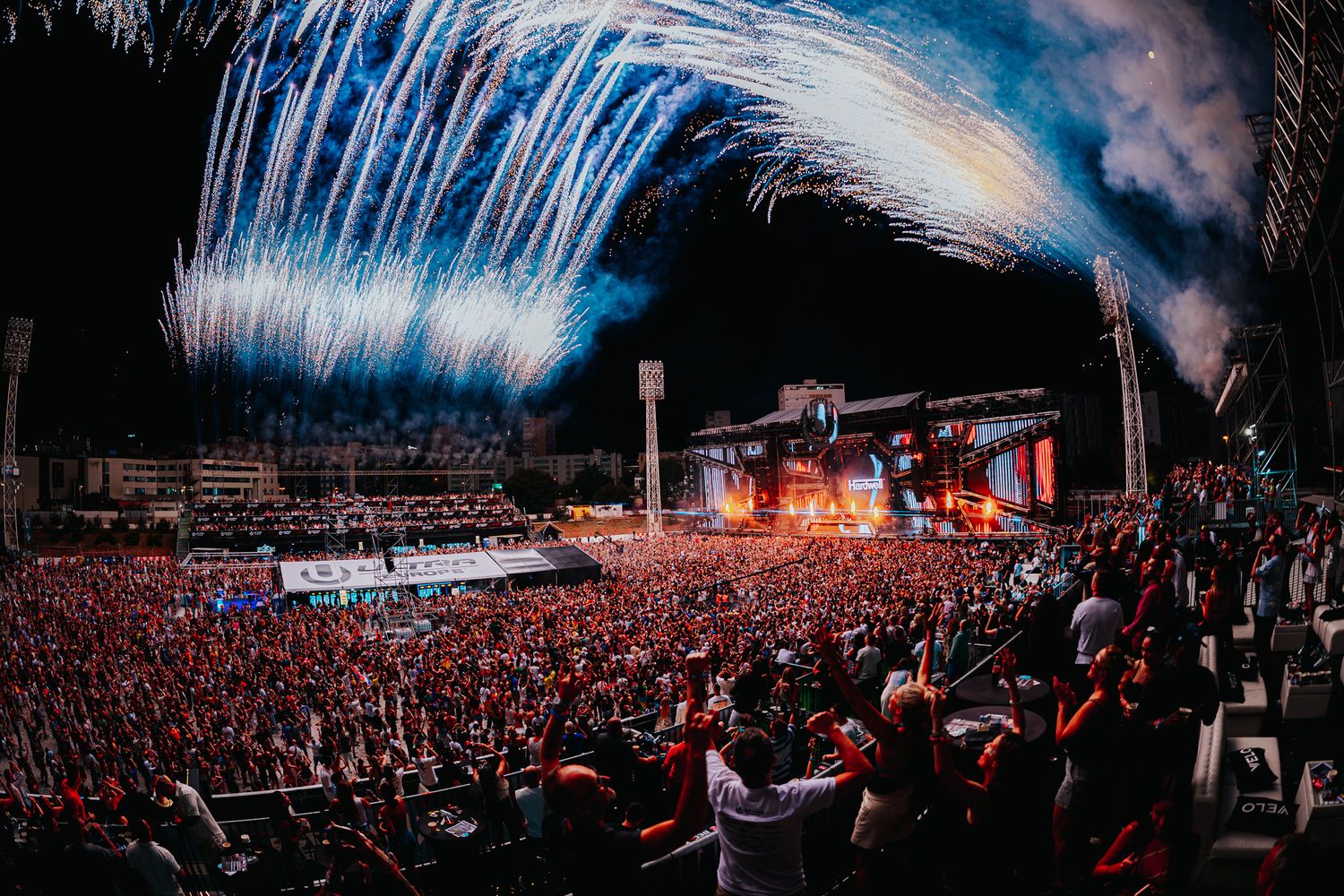 Massive outdoor concert with huge crowd, vibrant blue fireworks lighting the night sky above a brightly lit stage.