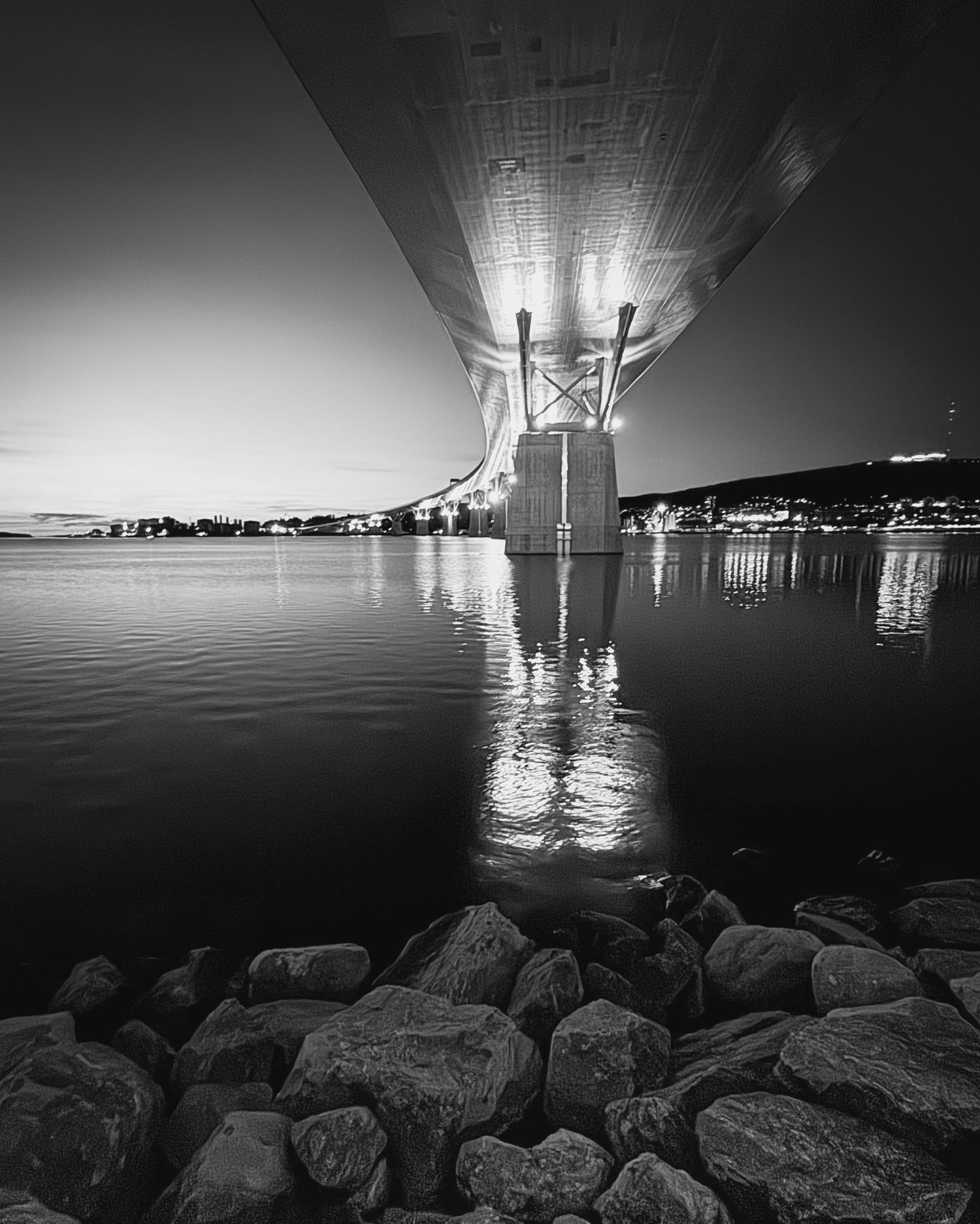 Black and white view under a bridge or pier reflecting on calm water with rocks in foreground.