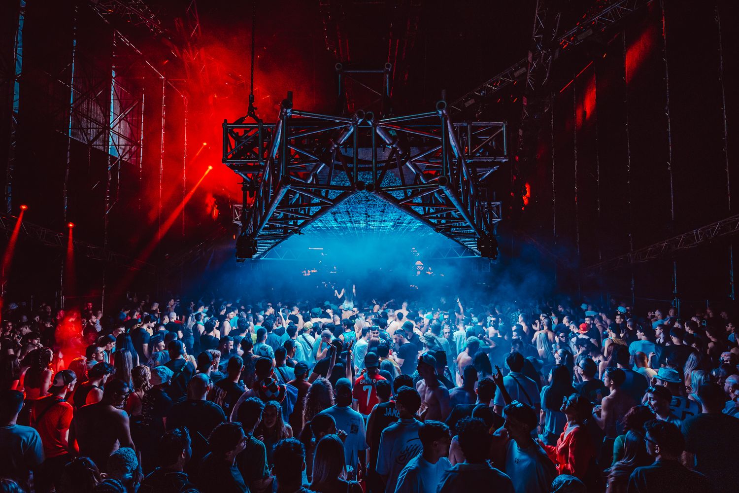 A large crowd dances under dramatic red and blue lights at an industrial-style indoor music festival venue.