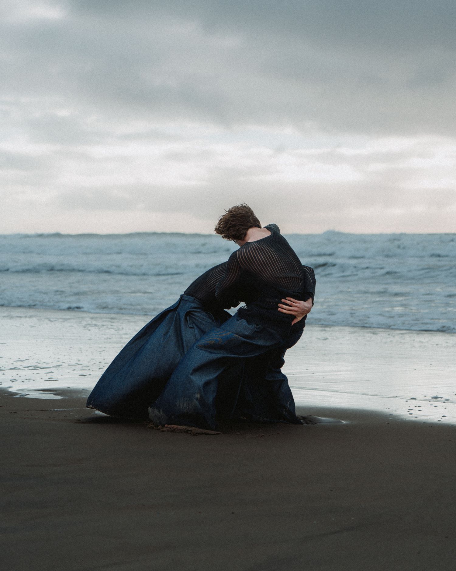 Duo de danse sur la plage, tissu en mouvement et étreinte intime sous un ciel dramatique.