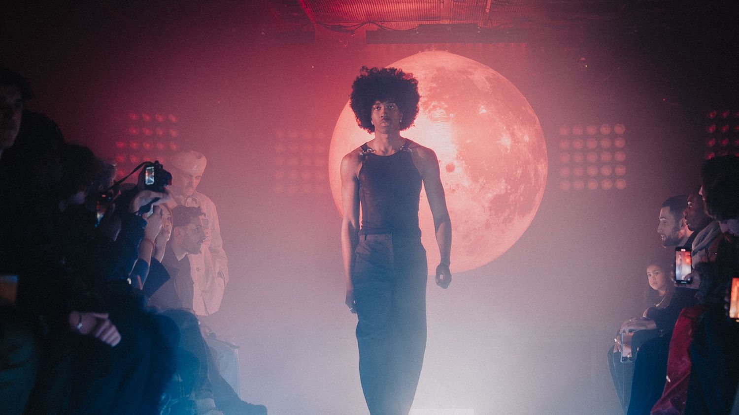 Défilé mode artistique, mannequin sur scène avec éclairage rouge et décor en lune, ambiance immersive.
