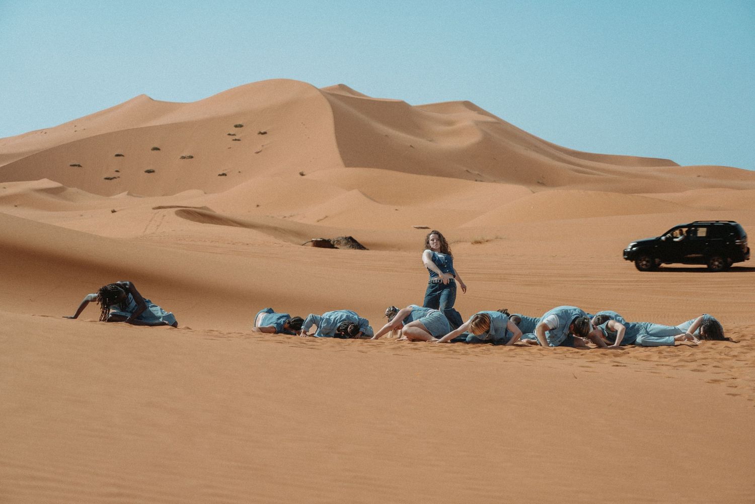 Une danseuse se tient debout face au ciel, entourée de corps allongés dans le sable, au cœur du désert de Merzouga