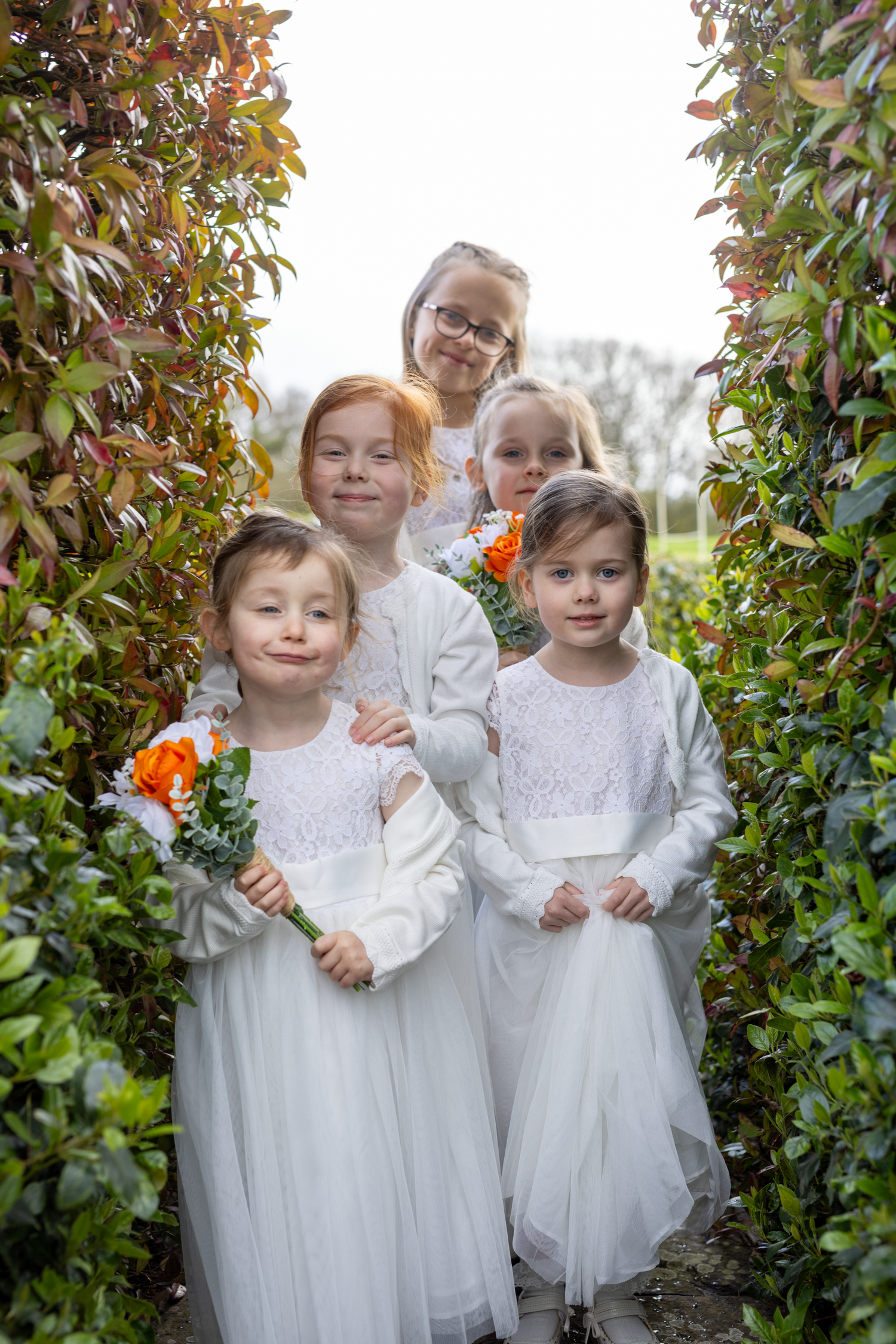 Five young flower girls in white dresses holding orange flowers posing in garden hedge pathway.