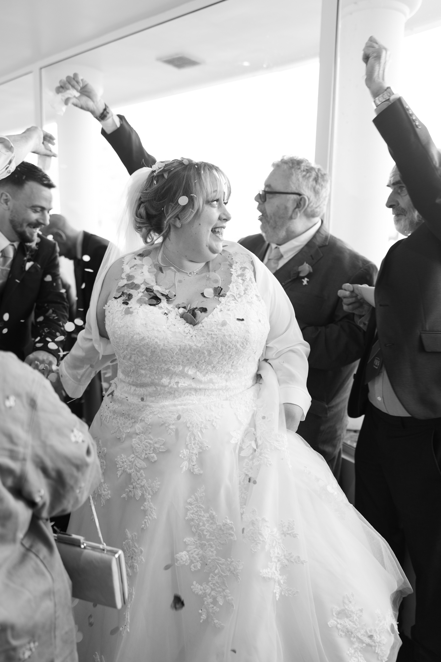 Happy bride in lace wedding gown celebrates with guests throwing confetti in black and white photo.
