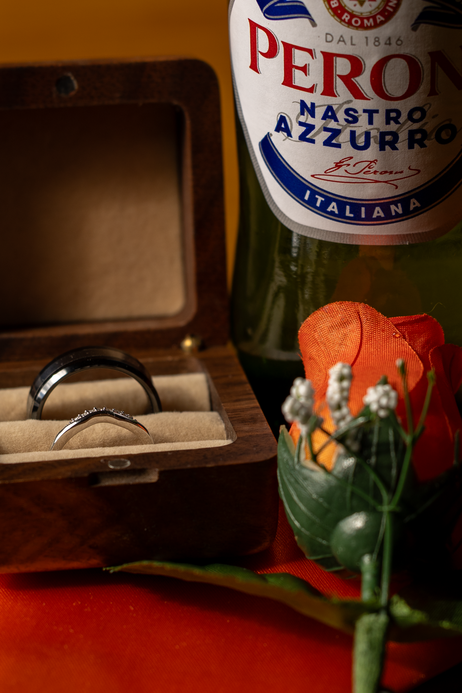Peroni Nastro Azzurro beer bottle beside a wooden ring box with wedding bands and a red rose.
