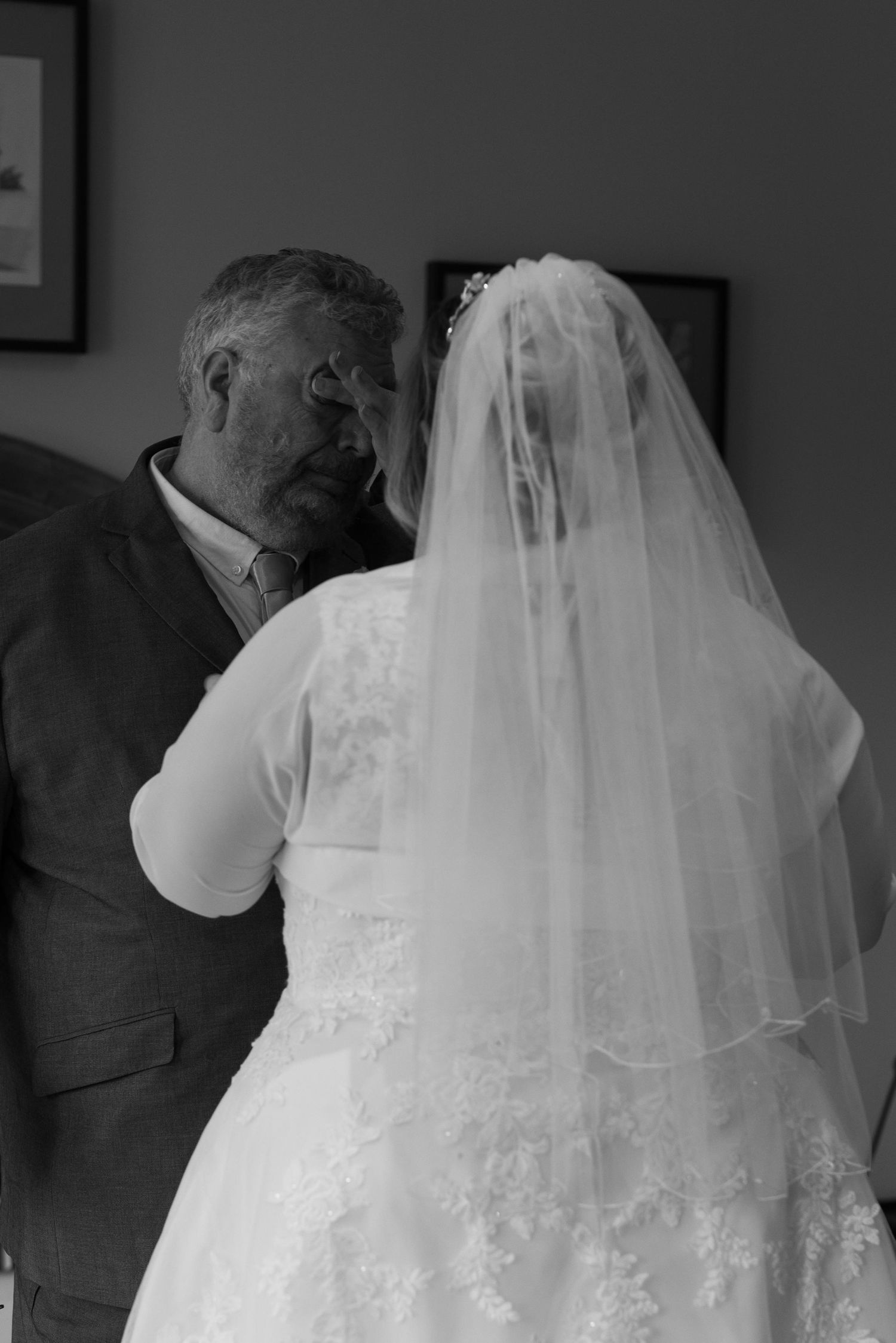 Black and white photo of a bride in lace gown and veil sharing a tender moment with an older man in a suit.