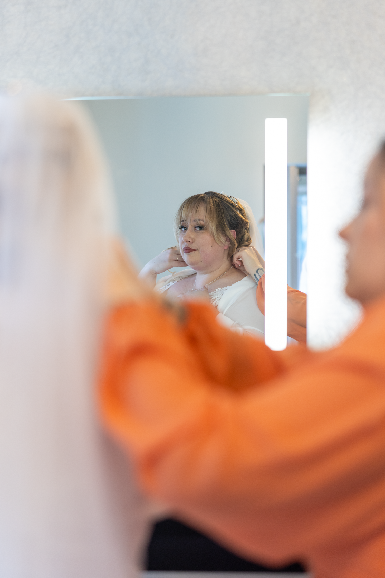 Bride getting ready, putting on earrings while looking in mirror with bridesmaid helping in orange top.