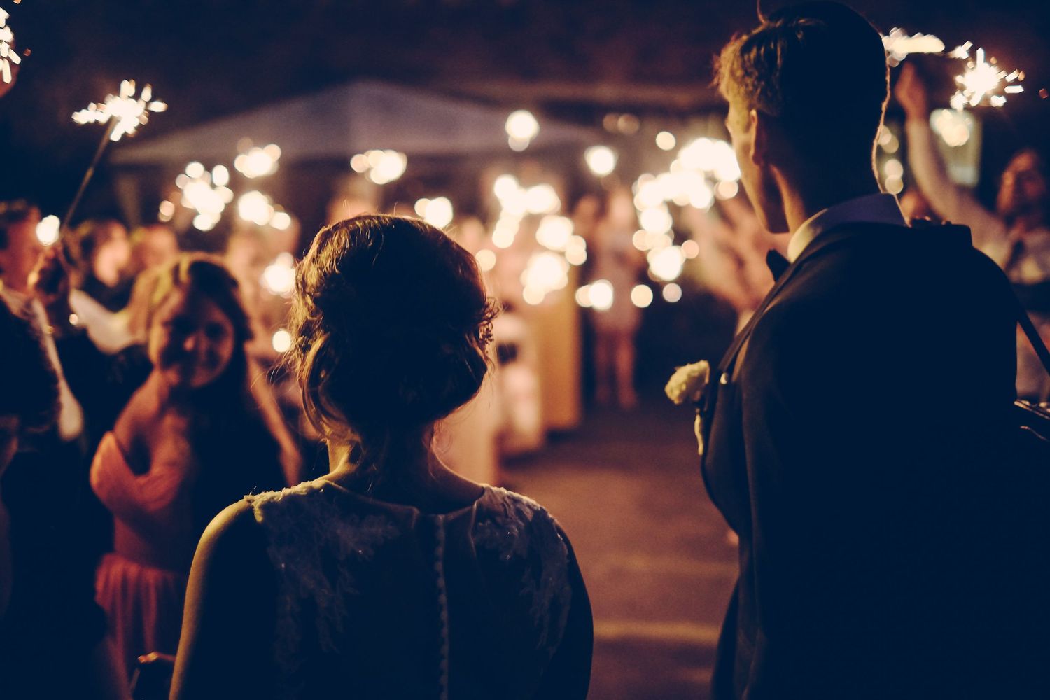 Bride and groom walk through sparkler tunnel at romantic outdoor wedding reception at night.