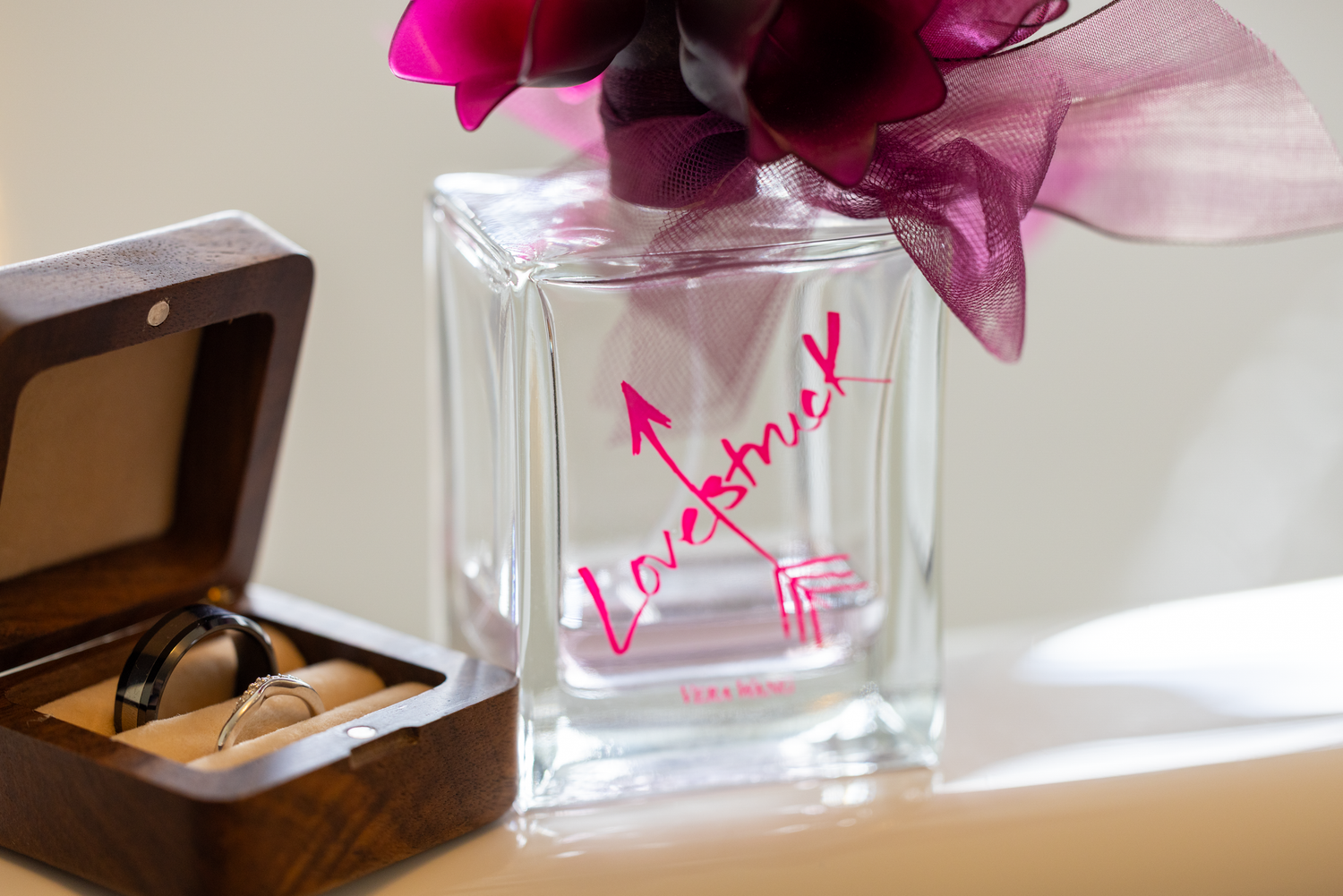 Wedding rings in wooden box next to Lovestruck perfume bottle and pink flowers on table.