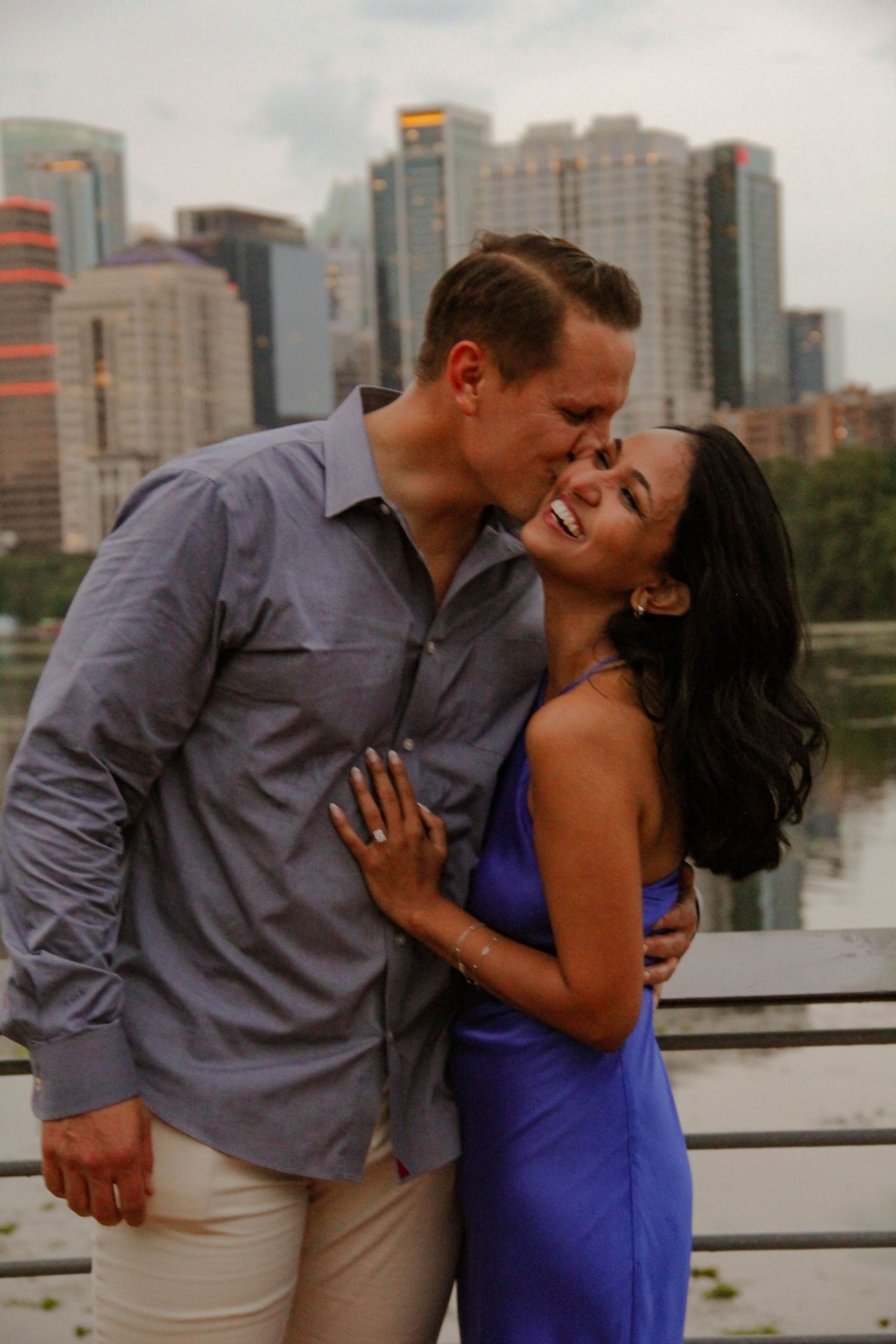 Happy newly engaged couple embracing and kissing with a city skyline reflected in water behind them.