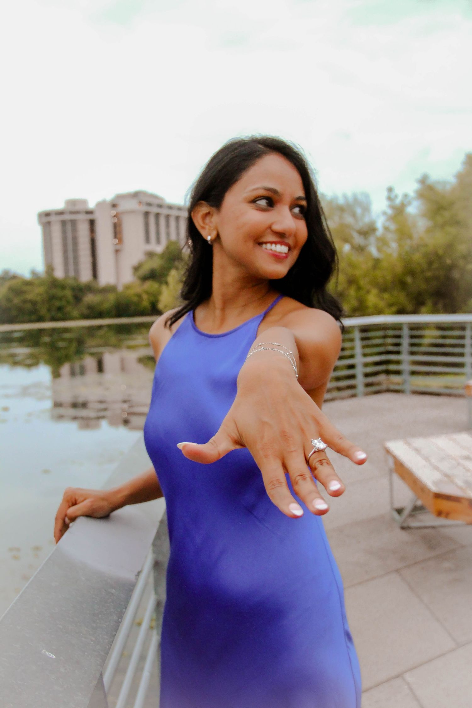 Joyful woman in blue dress proudly showing off her diamond engagement ring to the camera outdoors.