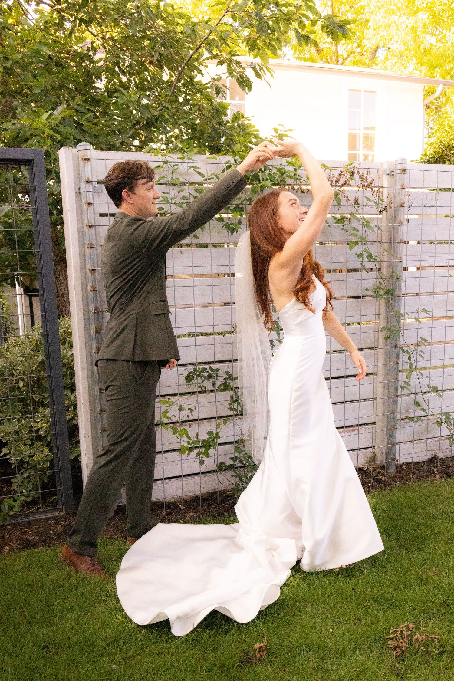 Groom in grey suit spinning bride in white backless wedding gown in garden setting.