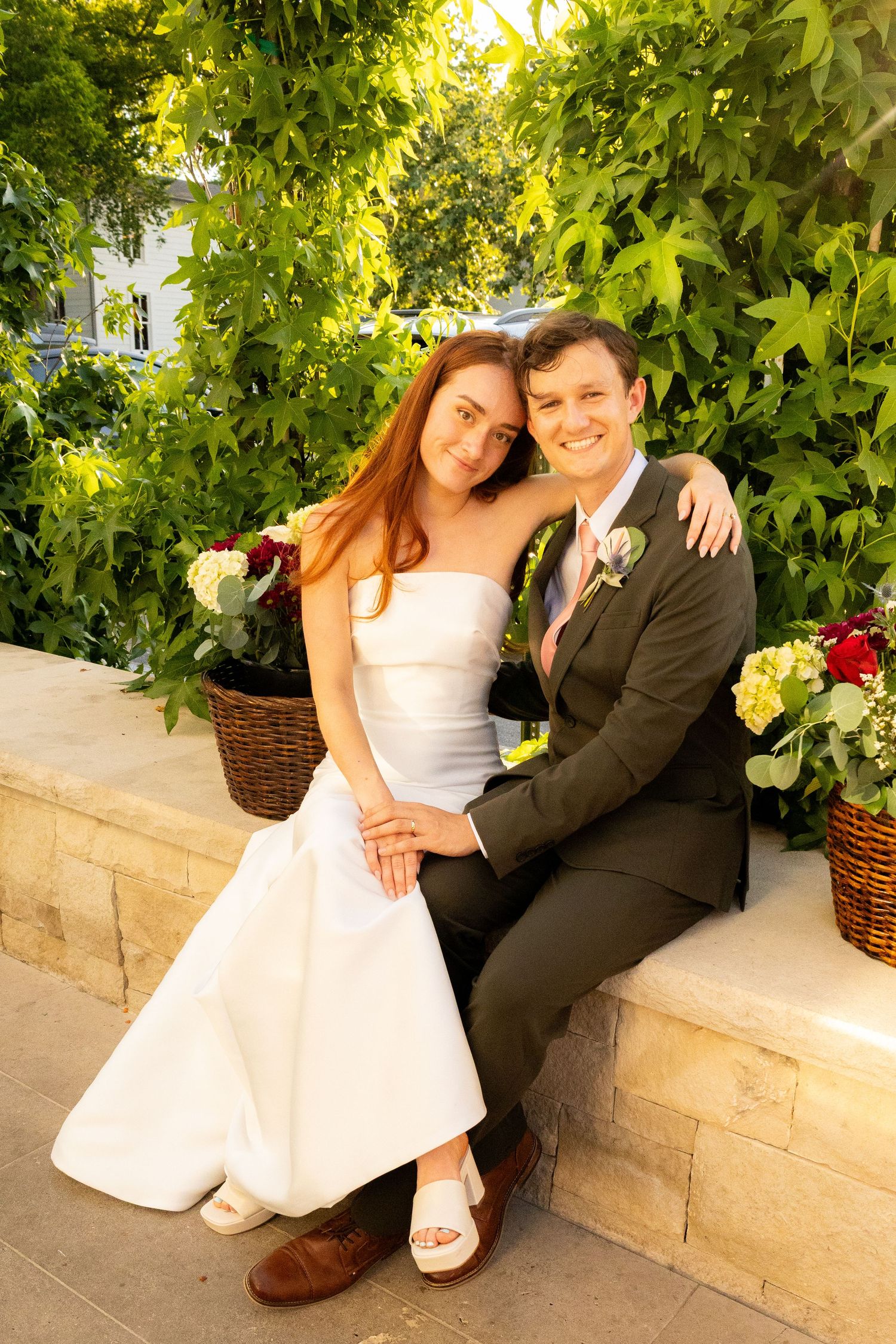 Bride and groom sitting together outdoors on stone steps, surrounded by lush greenery, smiling warmly on their wedding day.