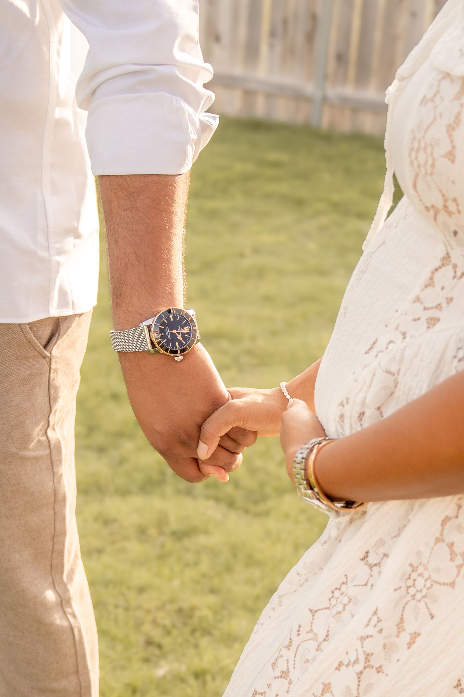 Couple holding hands outdoors, man wearing silver watch and white shirt, woman in white lace dress with gold bracelet.
