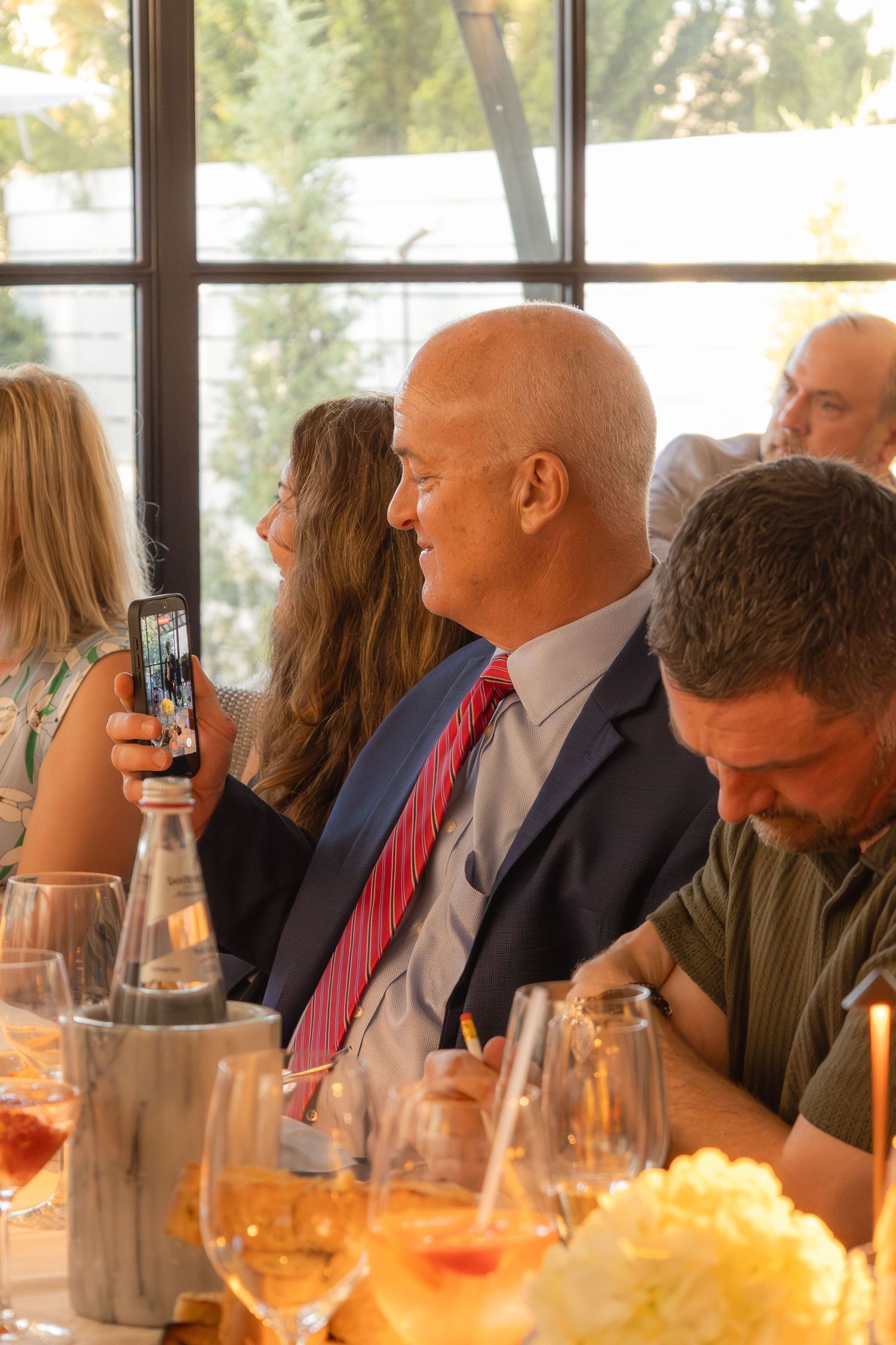 Bald man in navy suit and red tie smiling at a formal dinner event with wine glasses on the table.
