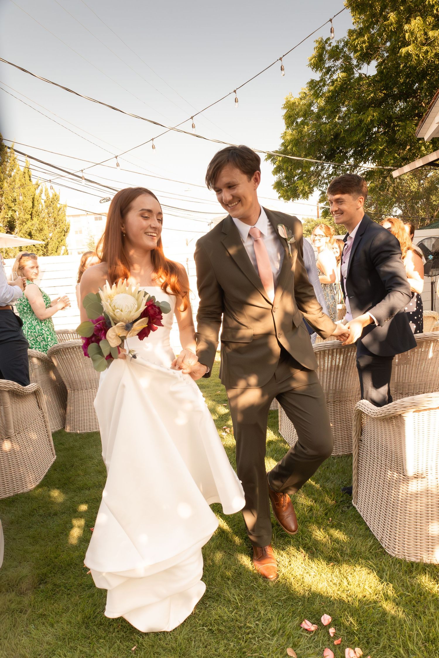 Bride and groom walk down outdoor aisle holding hands, smiling, with string lights and guests in background.