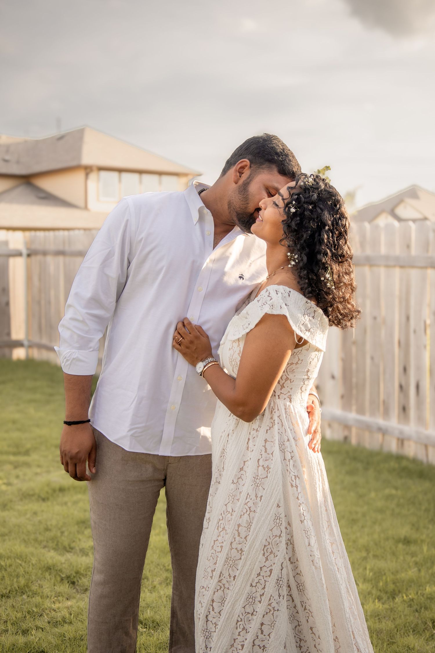 A couple shares a romantic kiss outdoors, dressed in white and beige, with a wooden fence and houses in the background.