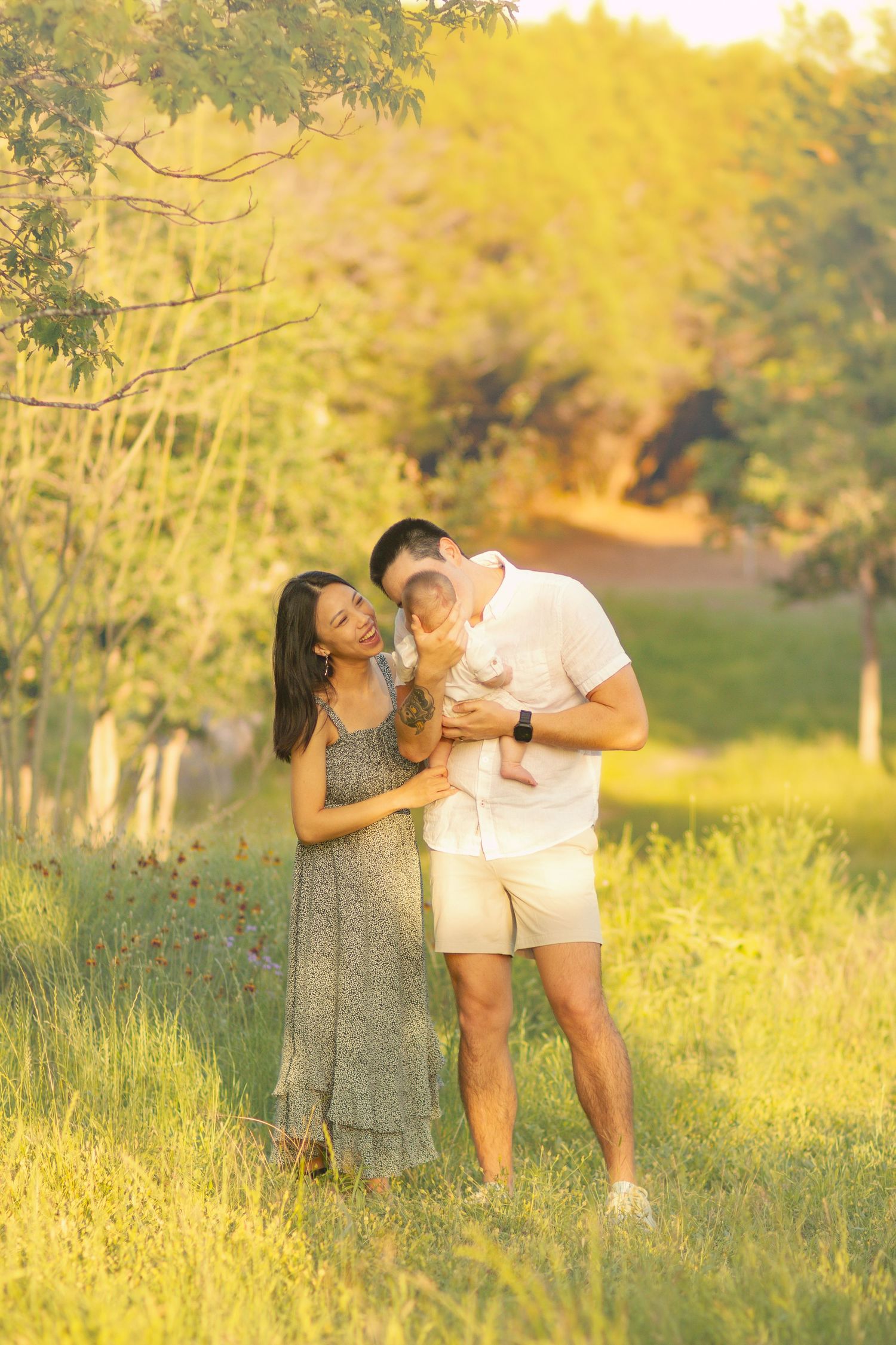 A happy young family with a baby enjoys a golden hour outdoor photo session in a lush green field.