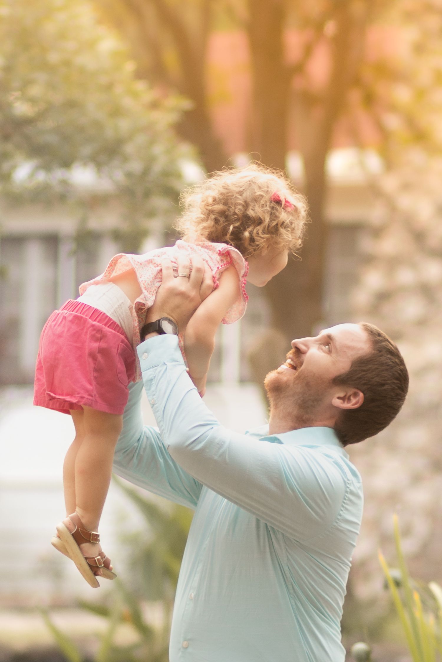 A smiling father lifts his curly-haired toddler girl in pink outfit overhead outdoors in a sunny backyard.