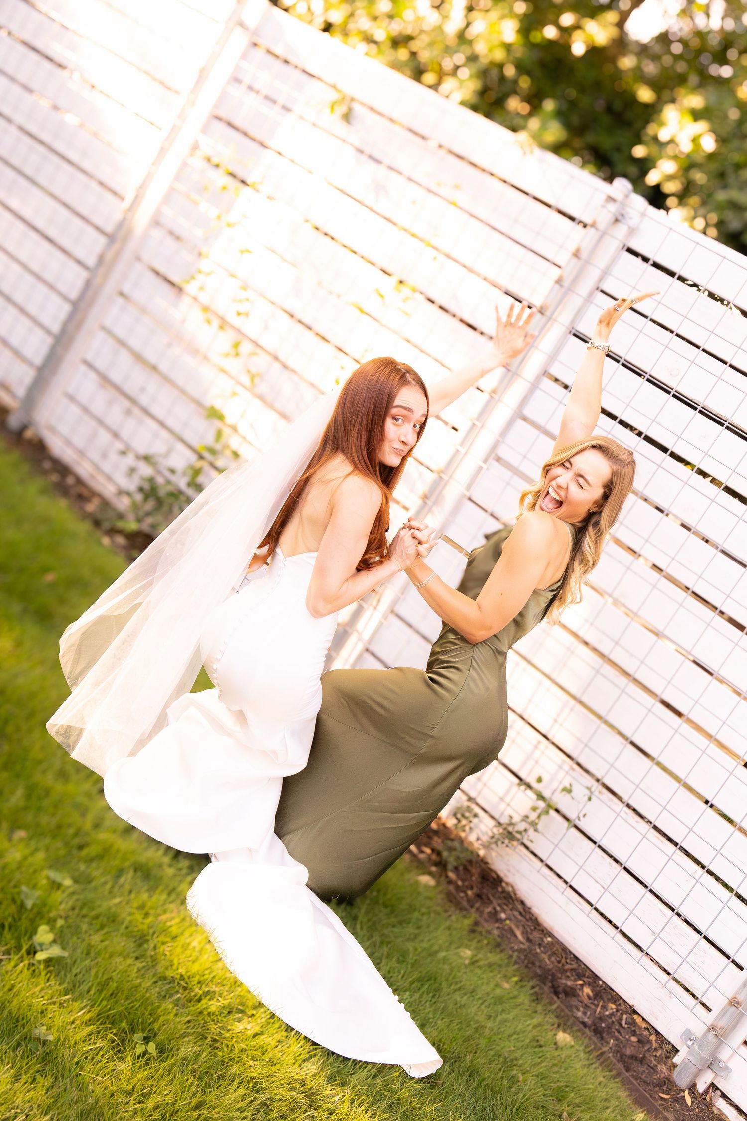 Bride in white gown laughs with bridesmaid in olive dress posing playfully by white fence.
