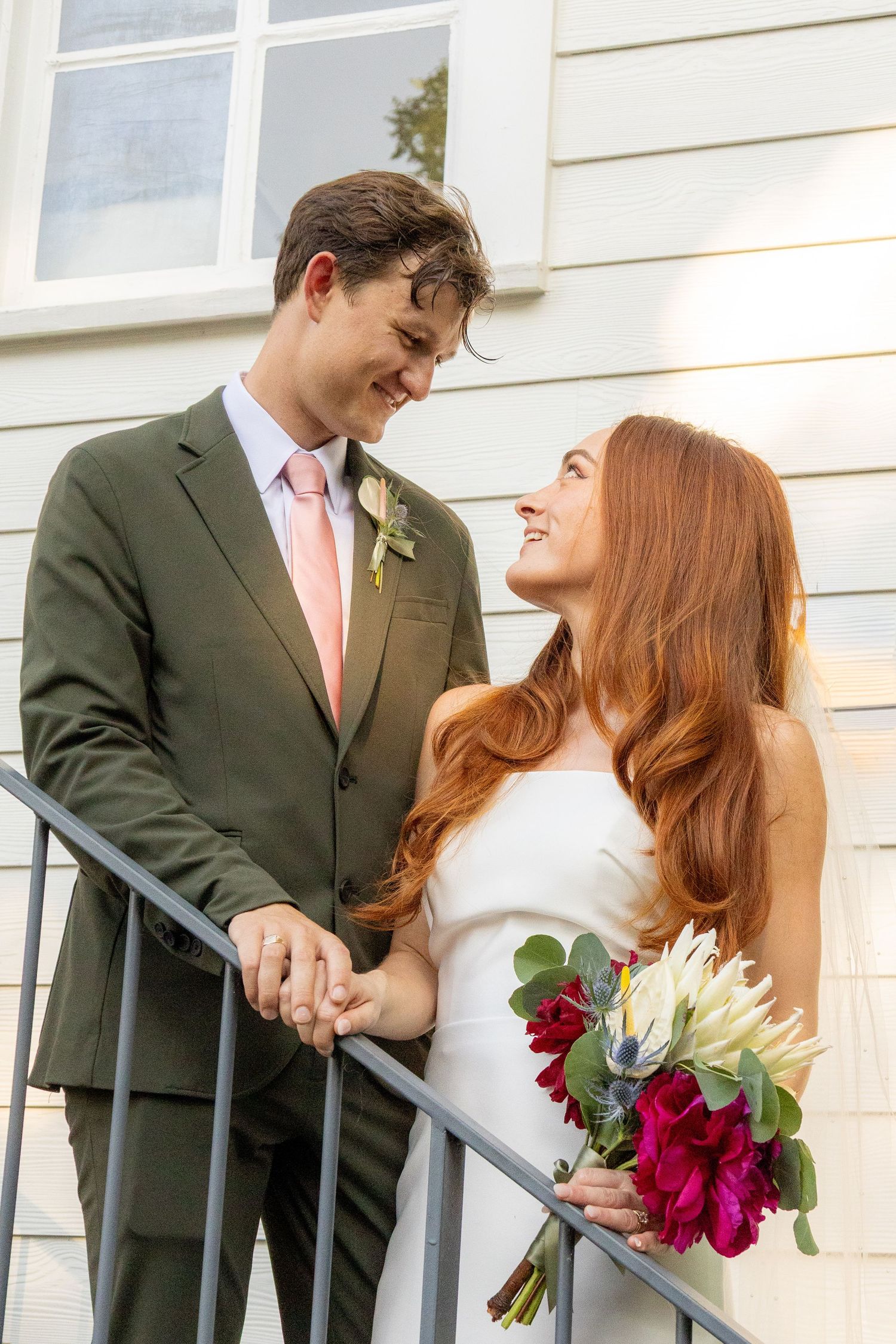 Smiling bride and groom holding hands on steps, bride in white dress with bouquet, groom in olive suit with pink tie.