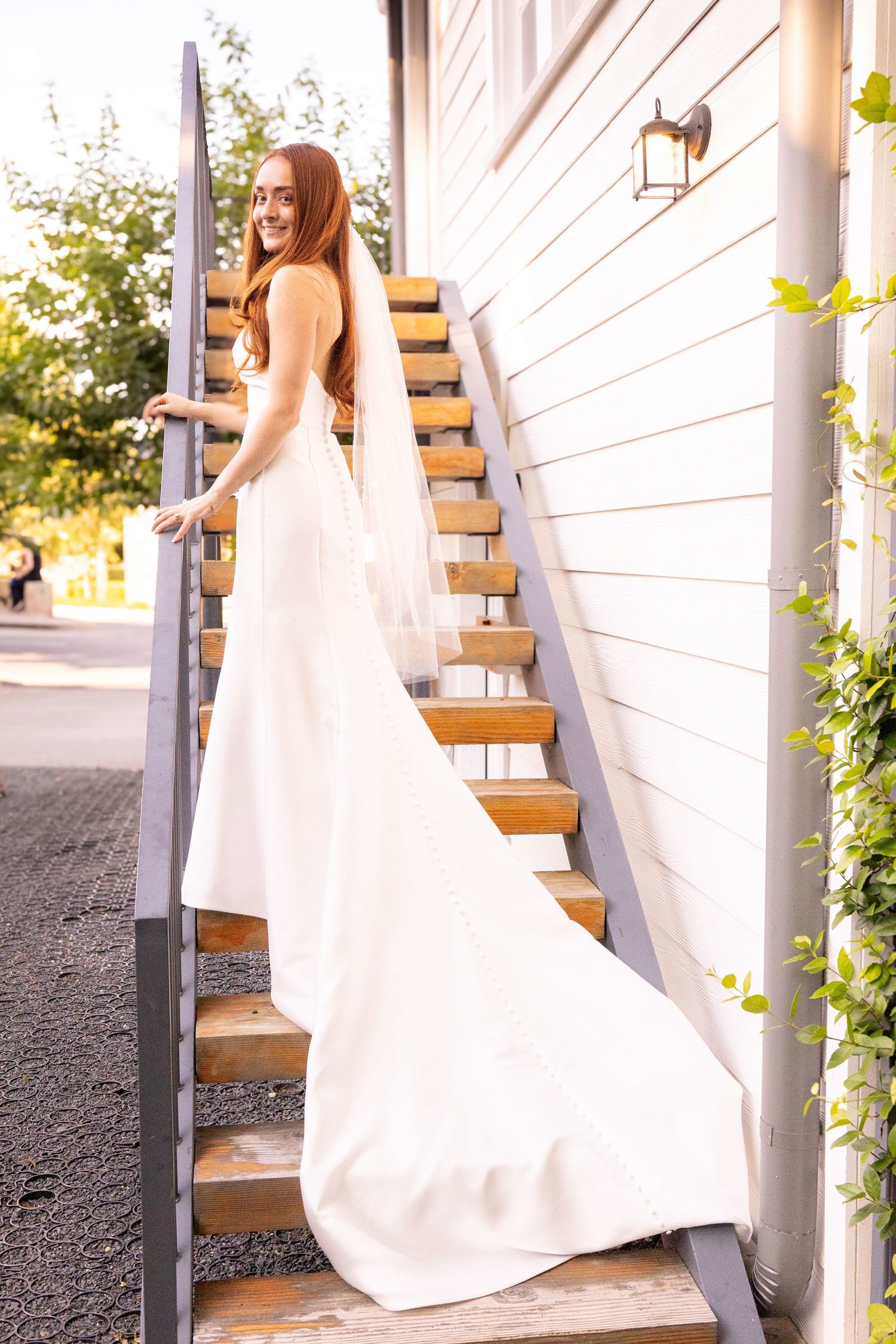 Smiling bride in elegant white gown with train stands on wooden outdoor staircase beside white building.