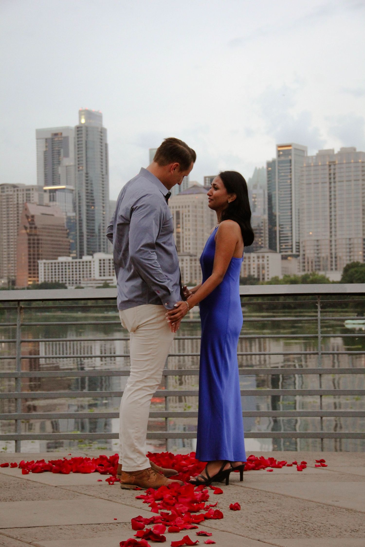 Engaged couple holding hands and facing each other surrounded by red rose petals and city skyline.