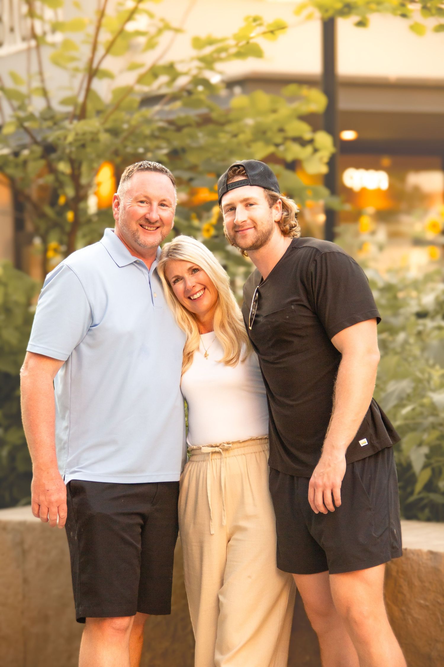 Three smiling adults posing together outdoors near greenery in casual summer attire.