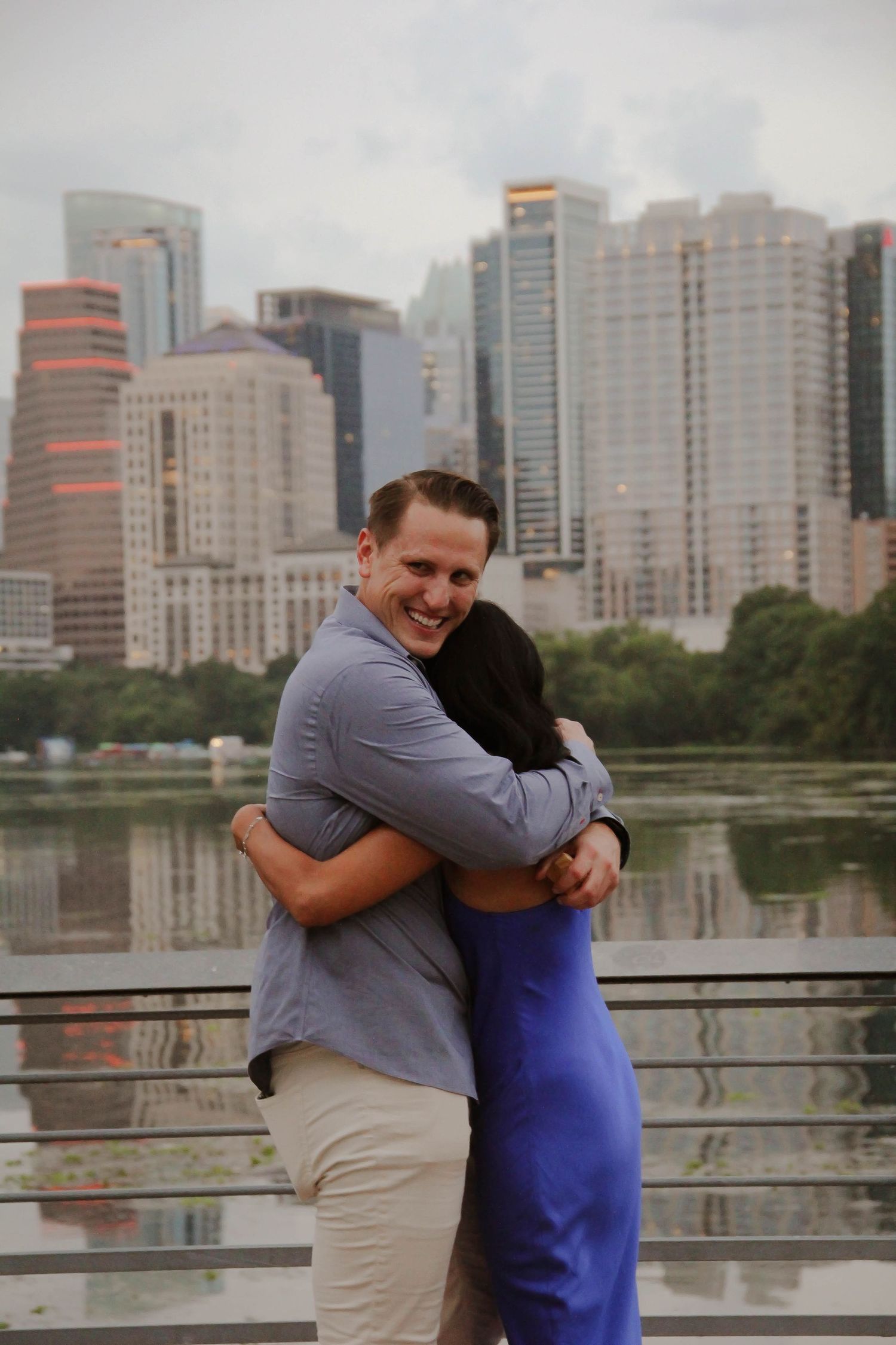 Couple hugging and smiling in front of a city skyline reflected in a calm lake on a cloudy day.