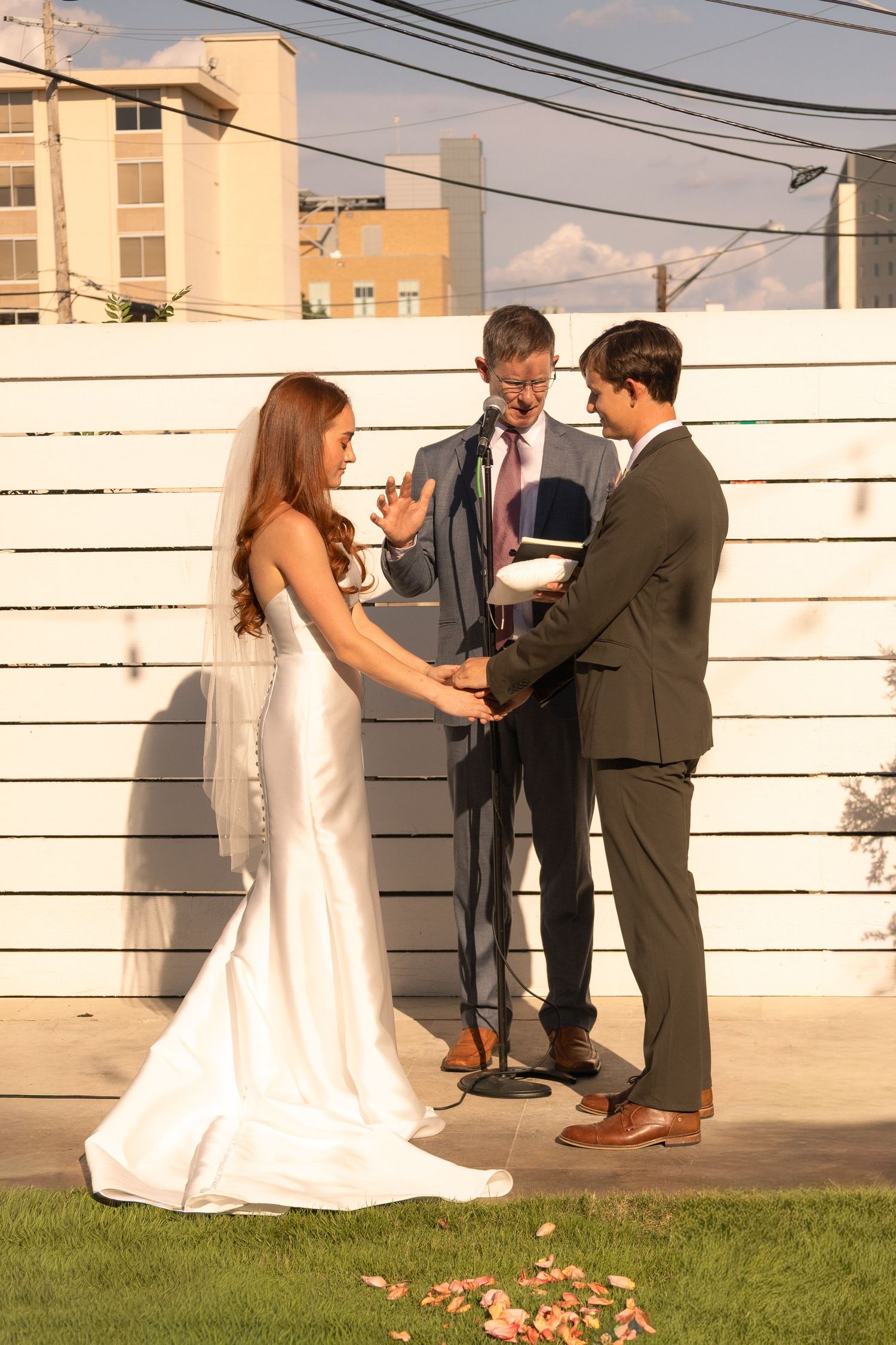 Outdoor wedding ceremony with bride in white gown and groom in suit exchanging vows with officiant at microphone.
