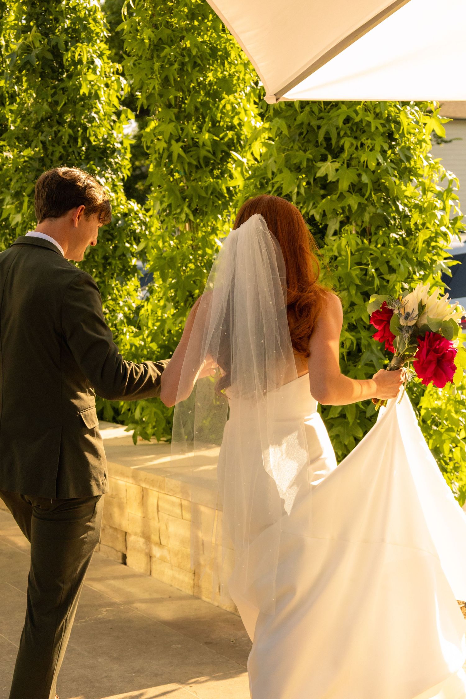 Bride and groom walking hand in hand outdoors, bride wearing white gown with veil holding red bouquet.