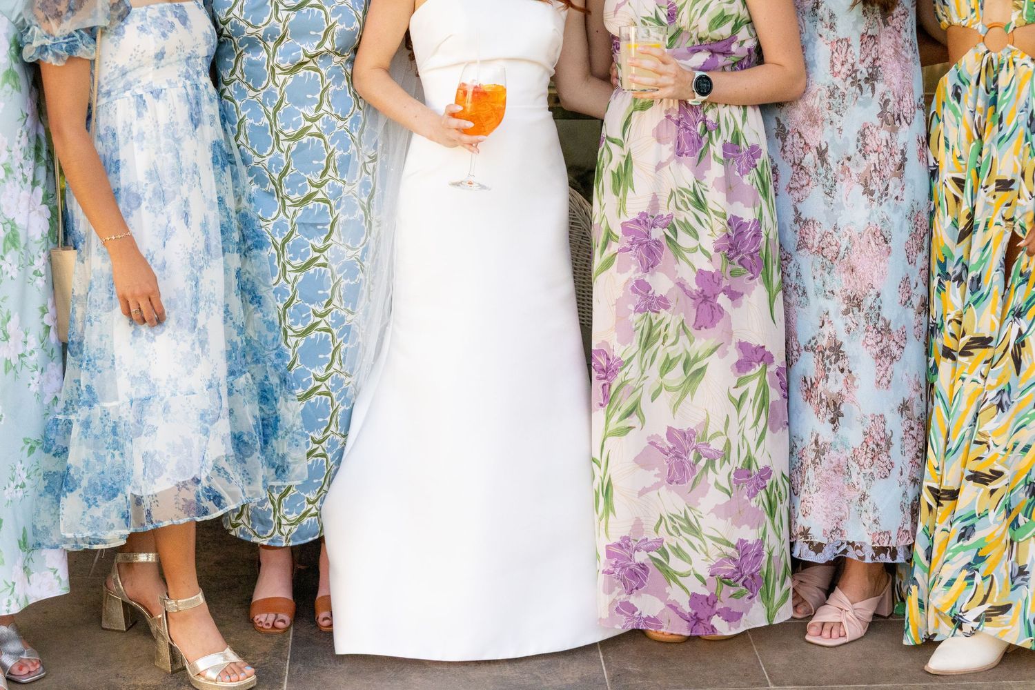 Bride in white gown surrounded by bridesmaids in colorful floral dresses holding drinks at a wedding celebration.