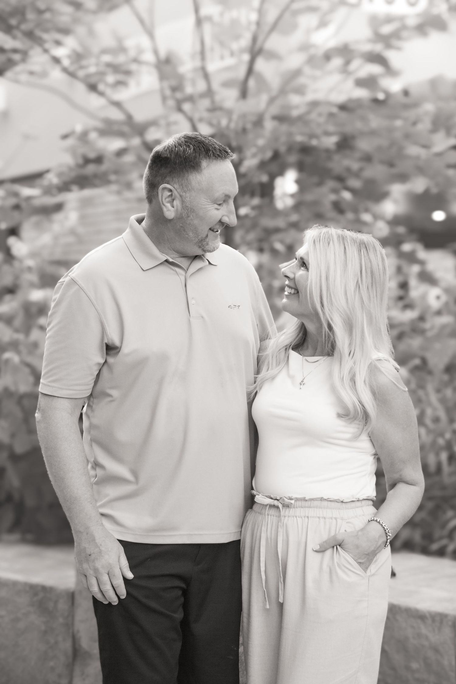 Black and white photo of a smiling couple standing together outdoors with lush green trees in the background.