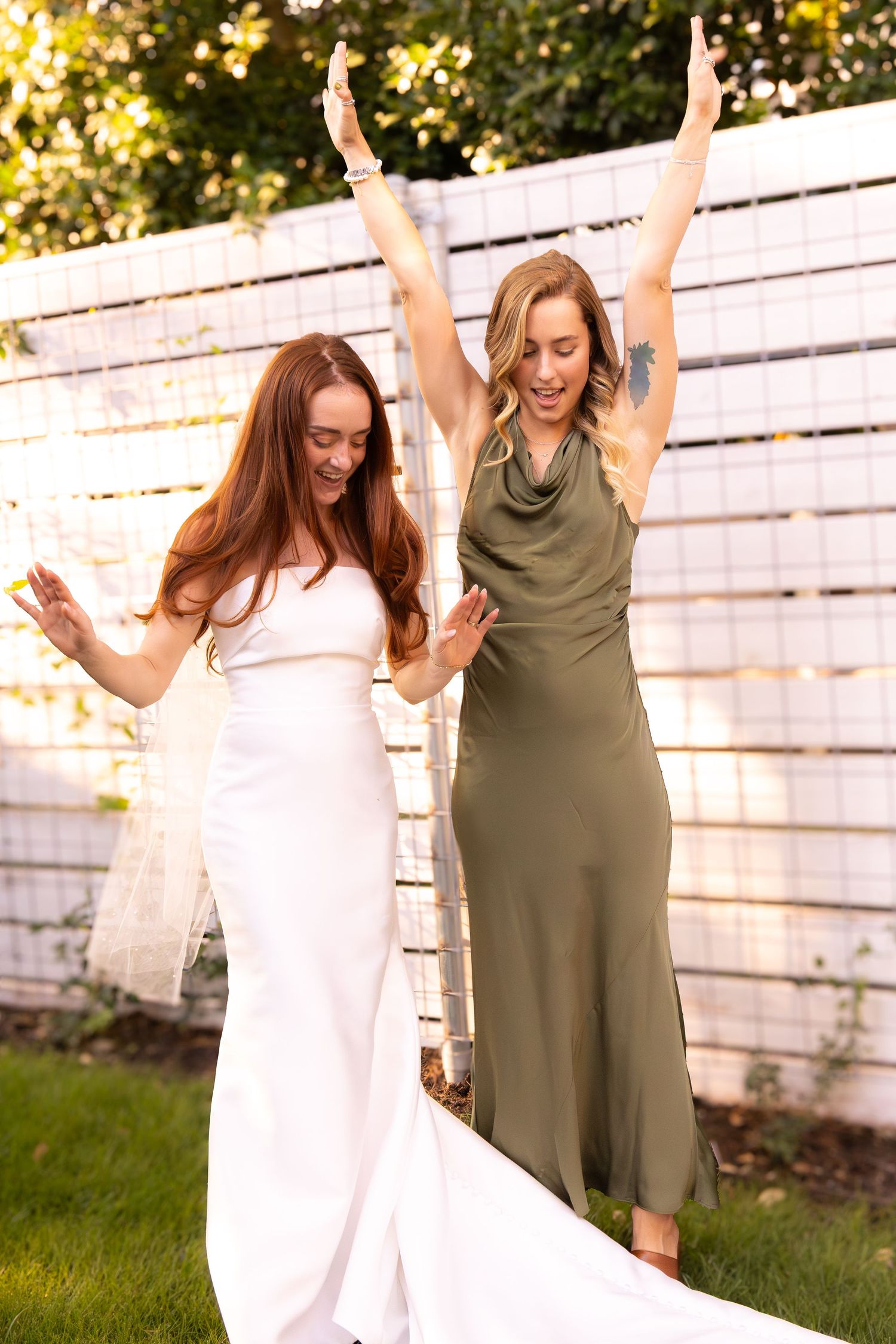 Bride in white gown and bridesmaid in olive dress celebrate joyfully with arms raised at outdoor wedding.