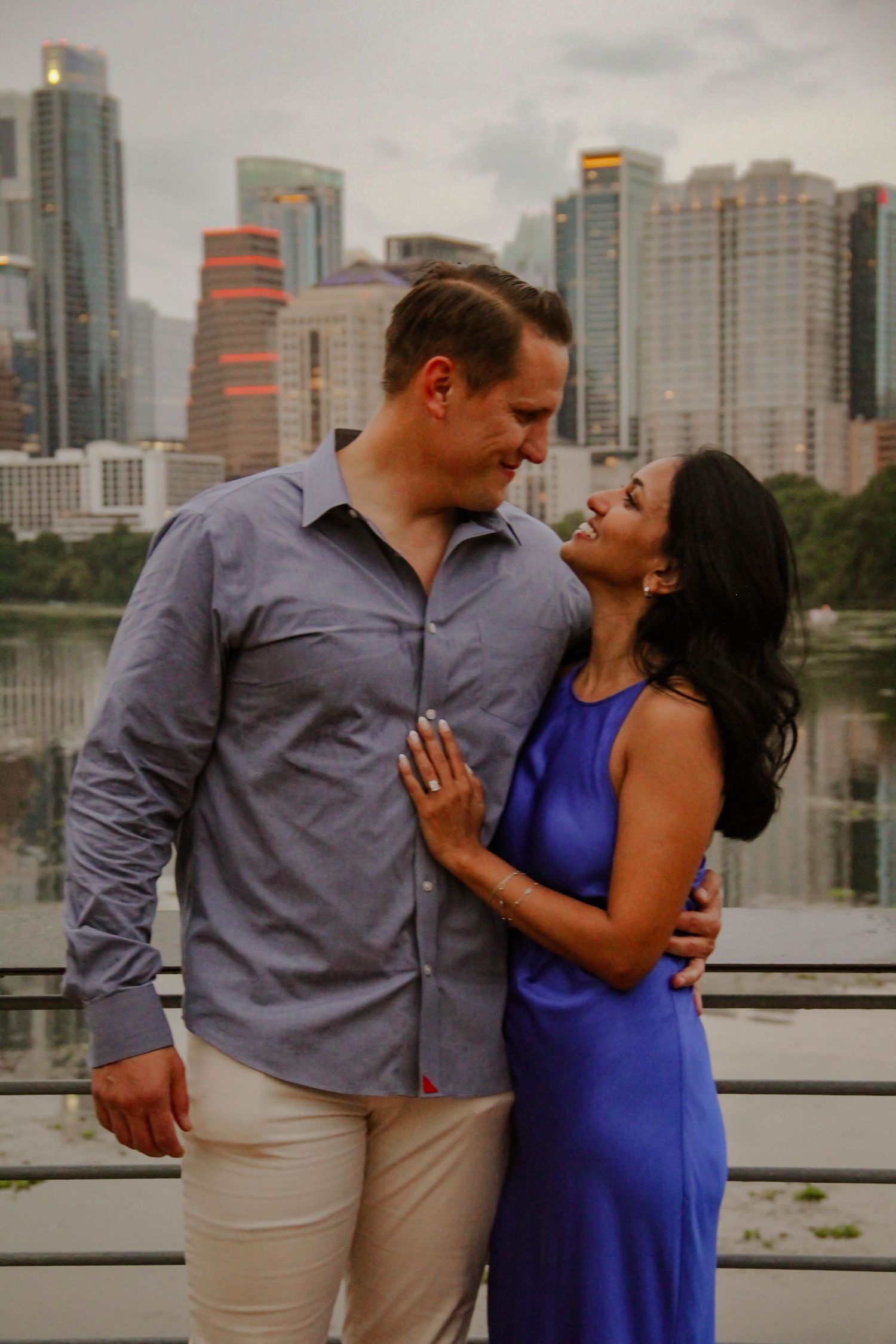 Couple sharing a romantic moment with a city skyline and lake backdrop, woman in blue dress and man in gray shirt.