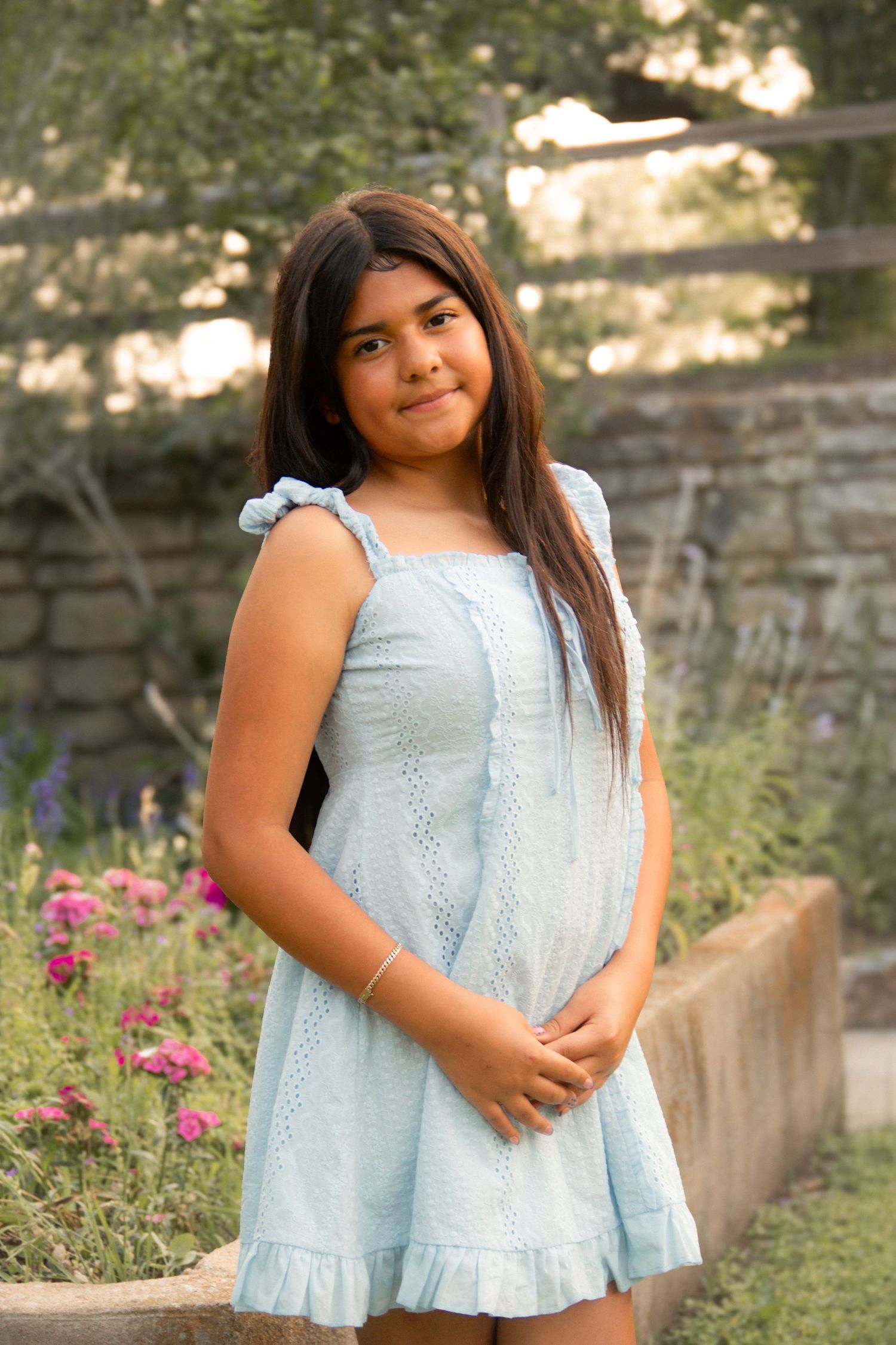Young girl in light blue sundress posing in garden with pink flowers.