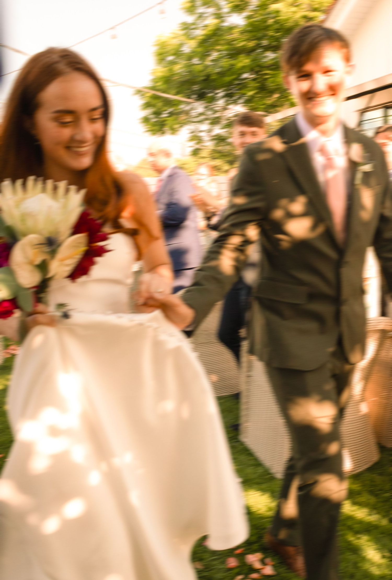 Smiling bride in white dress holding bouquet walks hand-in-hand with groom in gray suit at outdoor wedding ceremony.