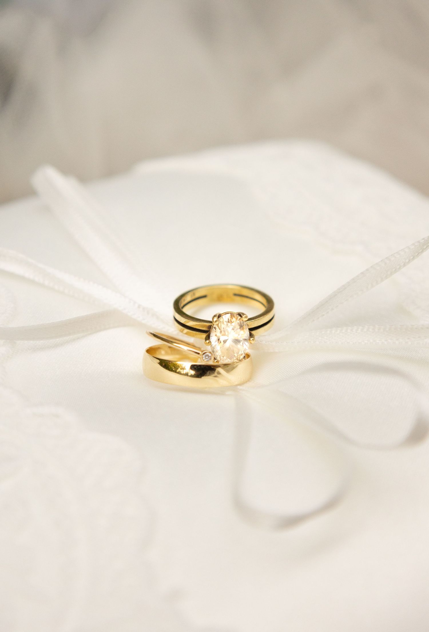 Gold wedding band set with oval diamond engagement ring displayed on white satin ring pillow with ribbon.