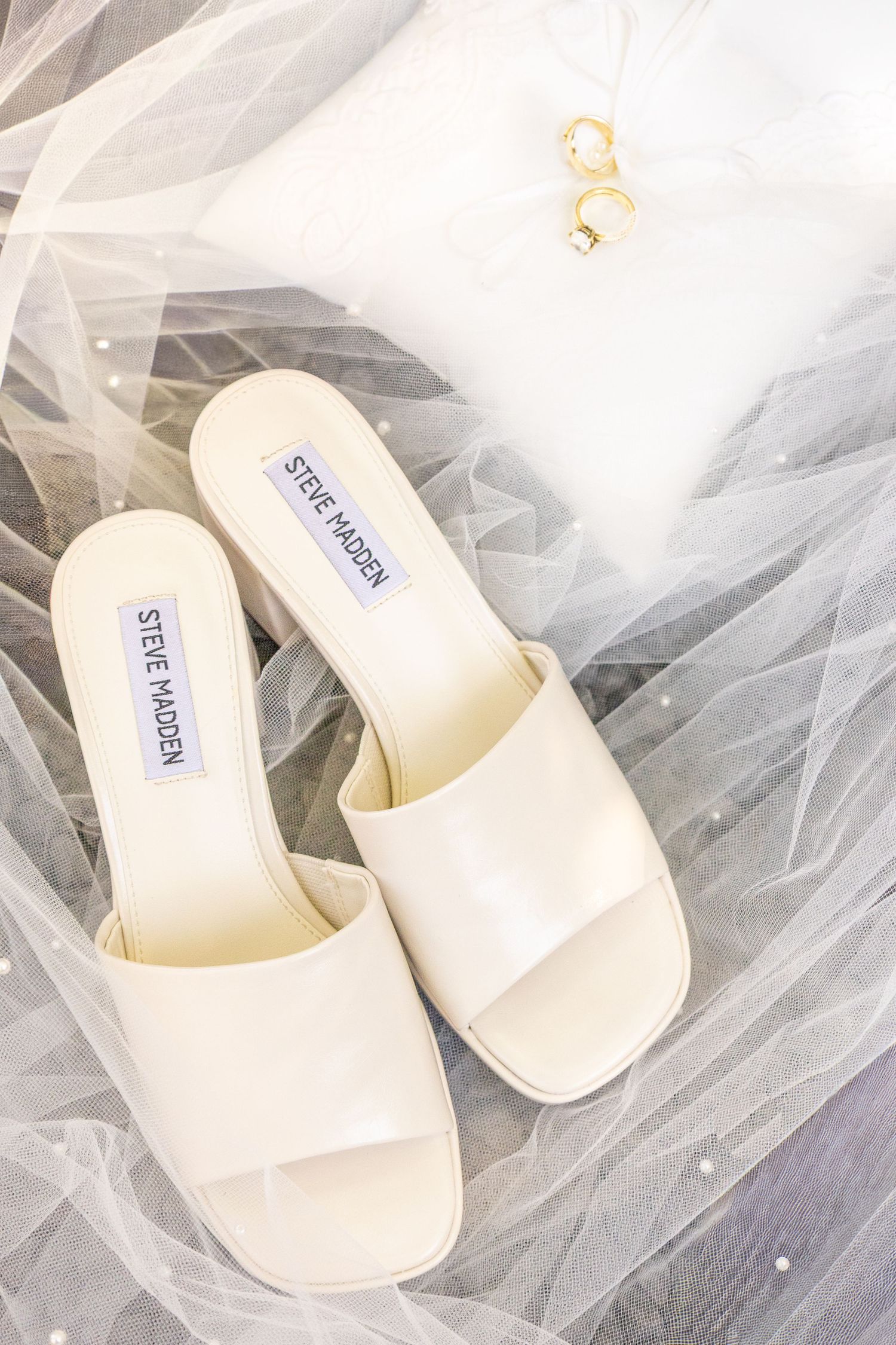 Steve Madden ivory block heel slide sandals displayed on white pillow with gold rings and bridal veil.