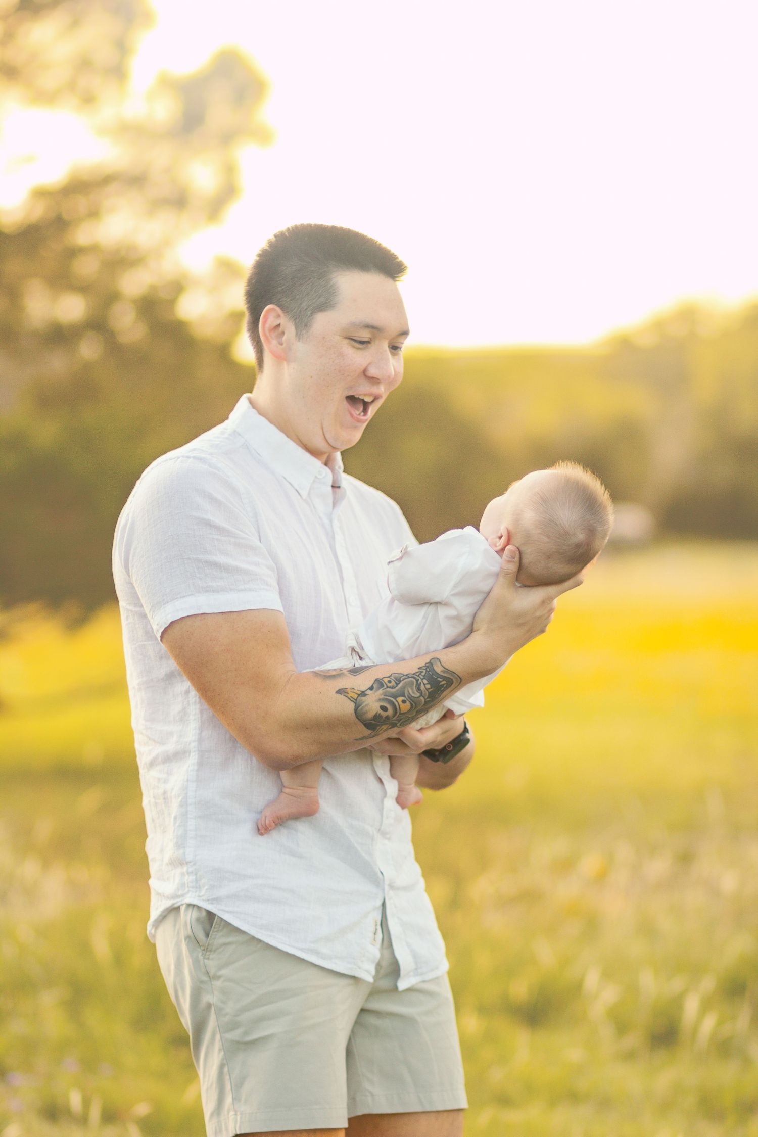 Father with tattoos holding newborn baby in sunny outdoor field, golden hour light, white shirt.