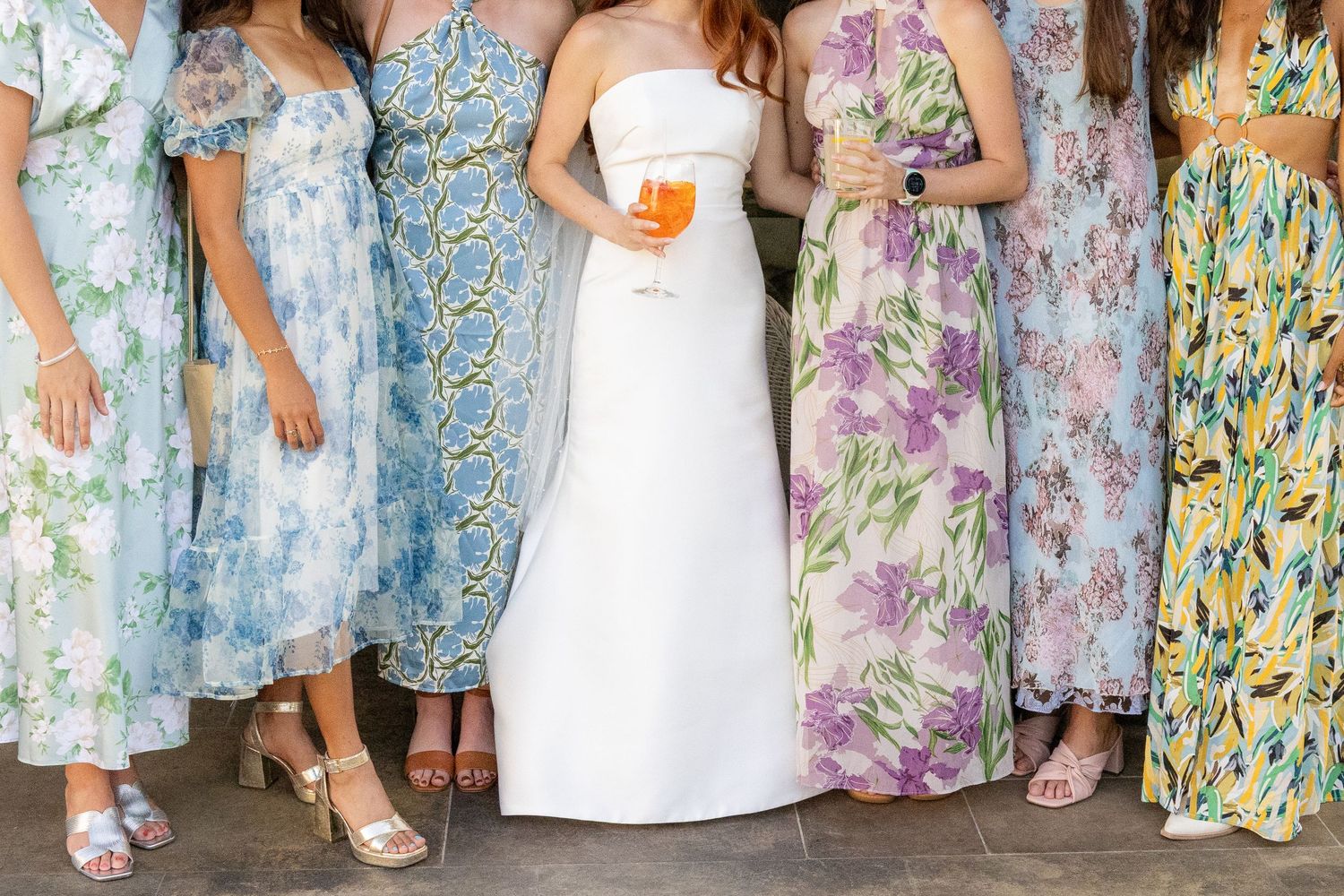 Bride in white strapless gown holding cocktail surrounded by bridesmaids in colorful floral maxi dresses.