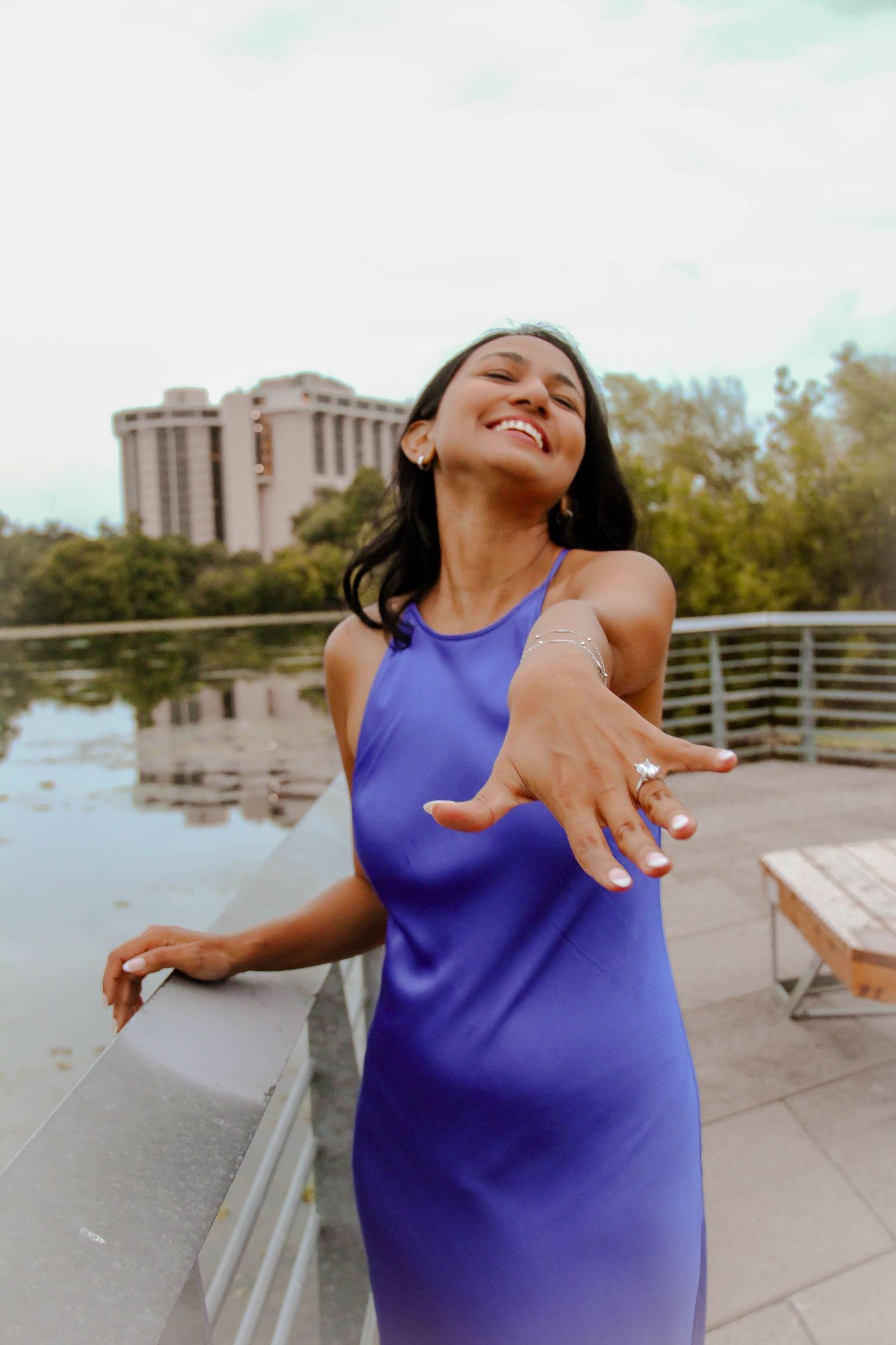 Joyful woman in a purple dress showing off her engagement ring while standing on an outdoor deck.