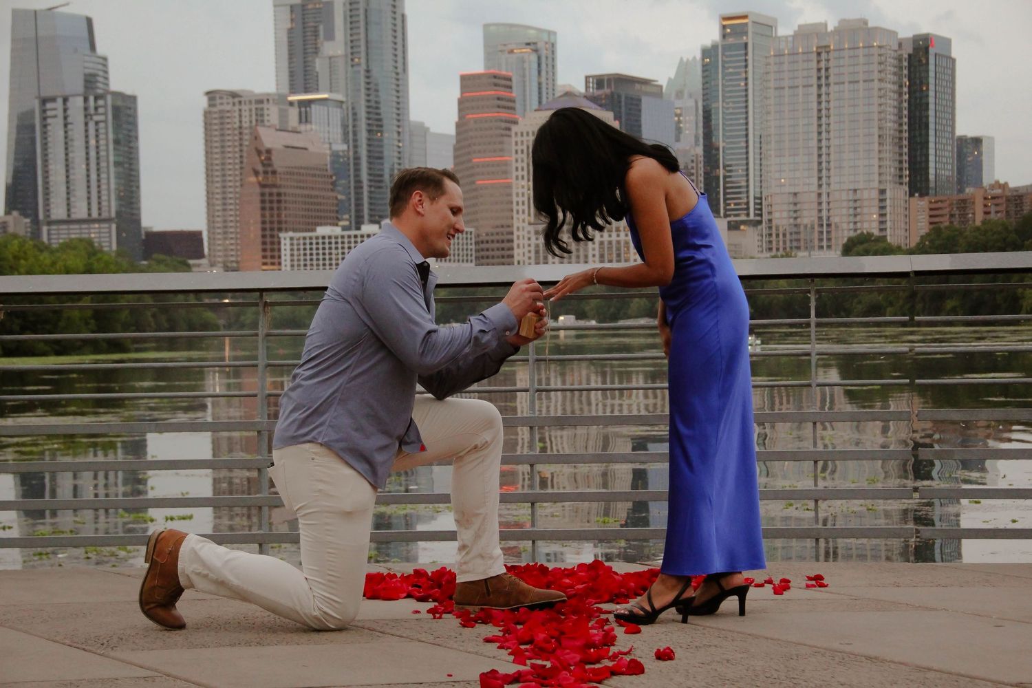Man proposes to woman on one knee with rose petals and city skyline backdrop.