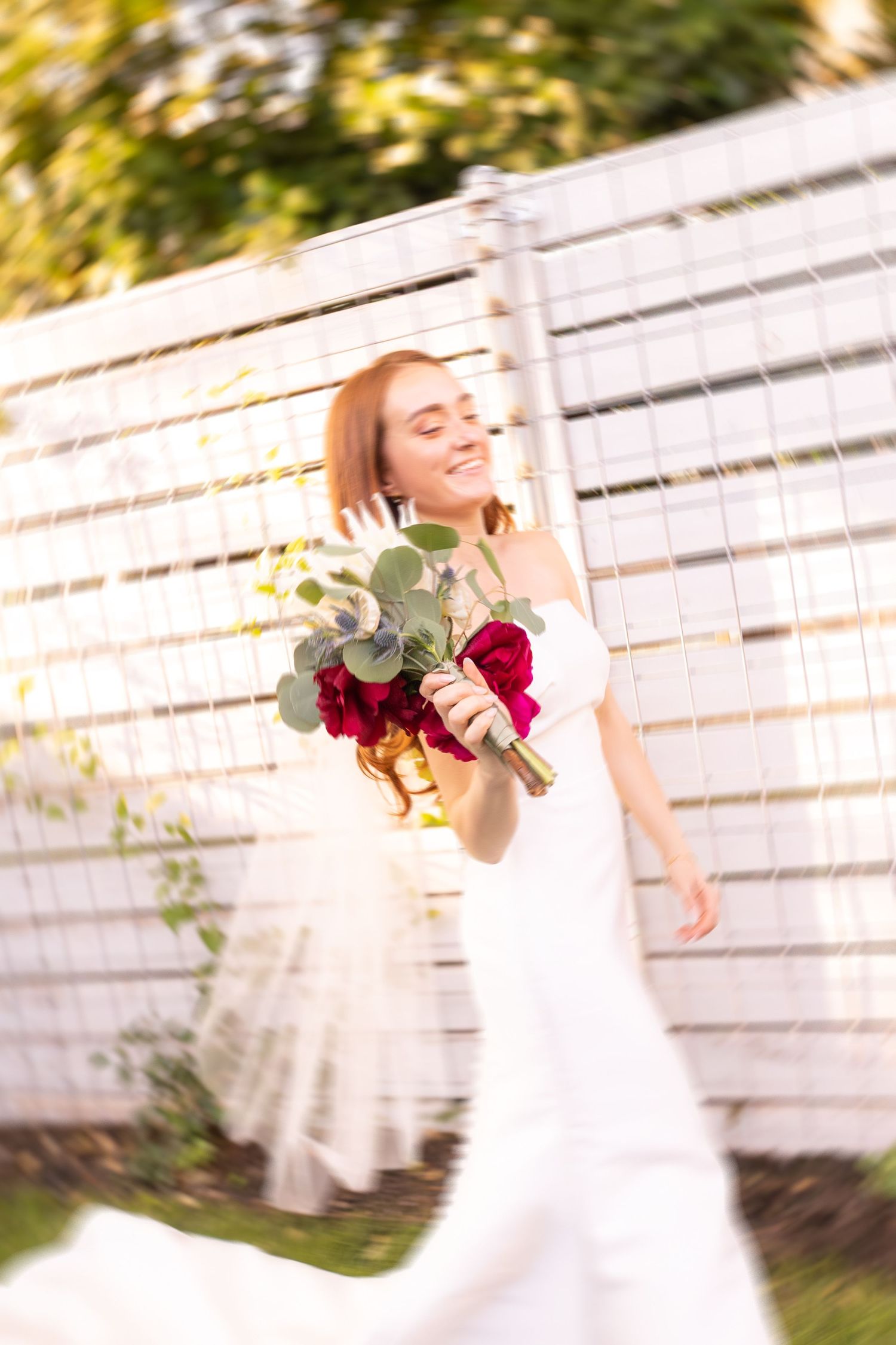 A joyful bride in a white gown twirls outdoors, holding a bouquet of red roses and eucalyptus near a white fence.