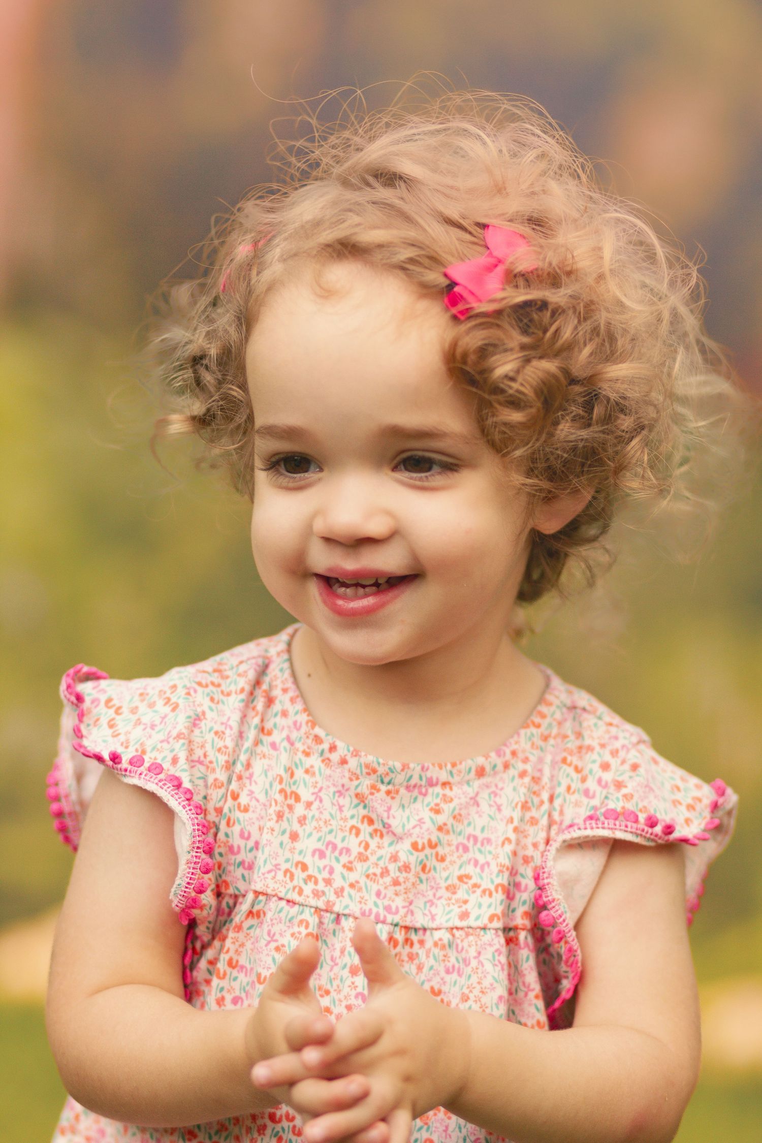 Smiling toddler girl with curly hair and pink bow wearing floral dress, clapping hands outdoors.