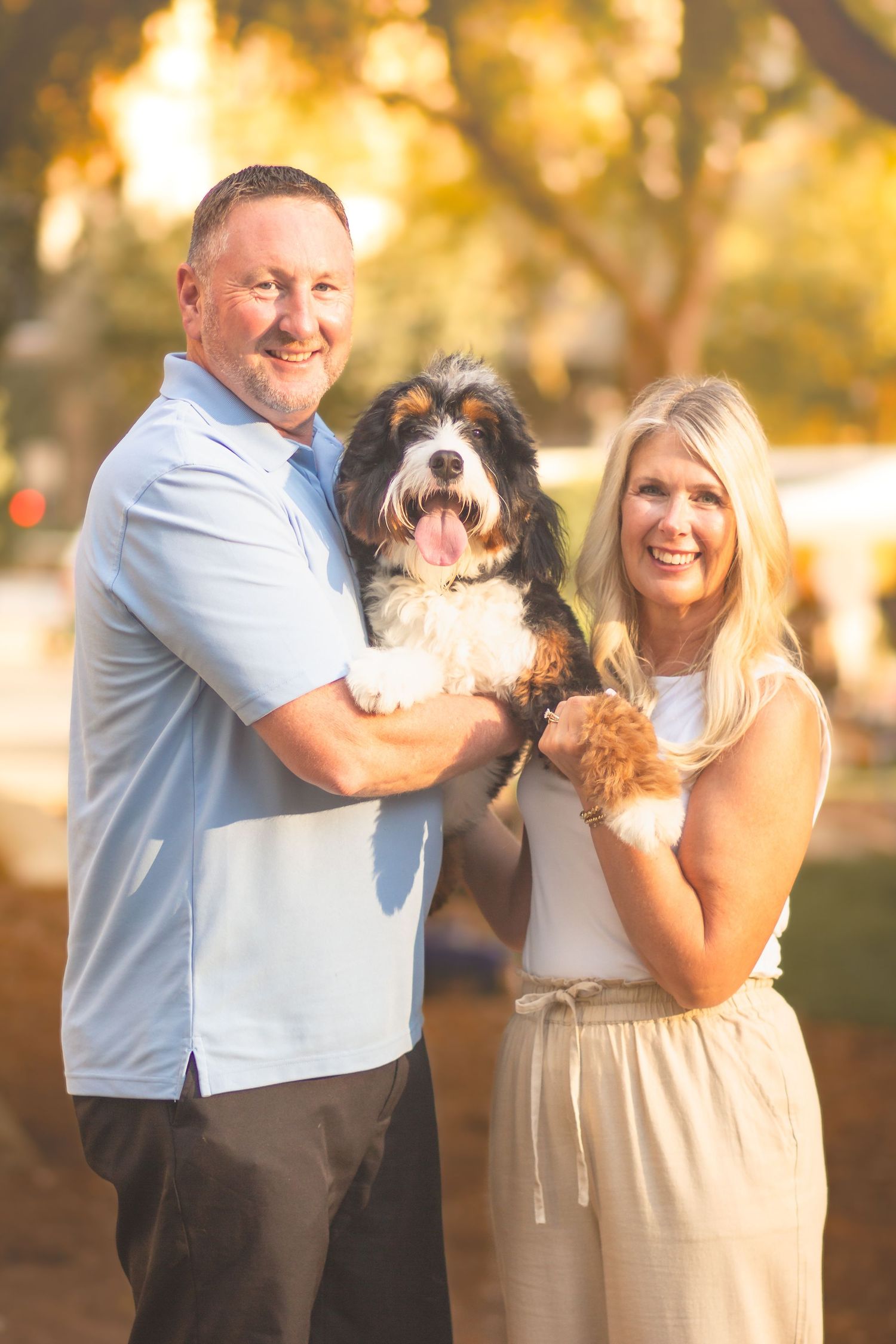 Smiling couple holding a fluffy Bernedoodle dog outdoors in warm golden sunlight.