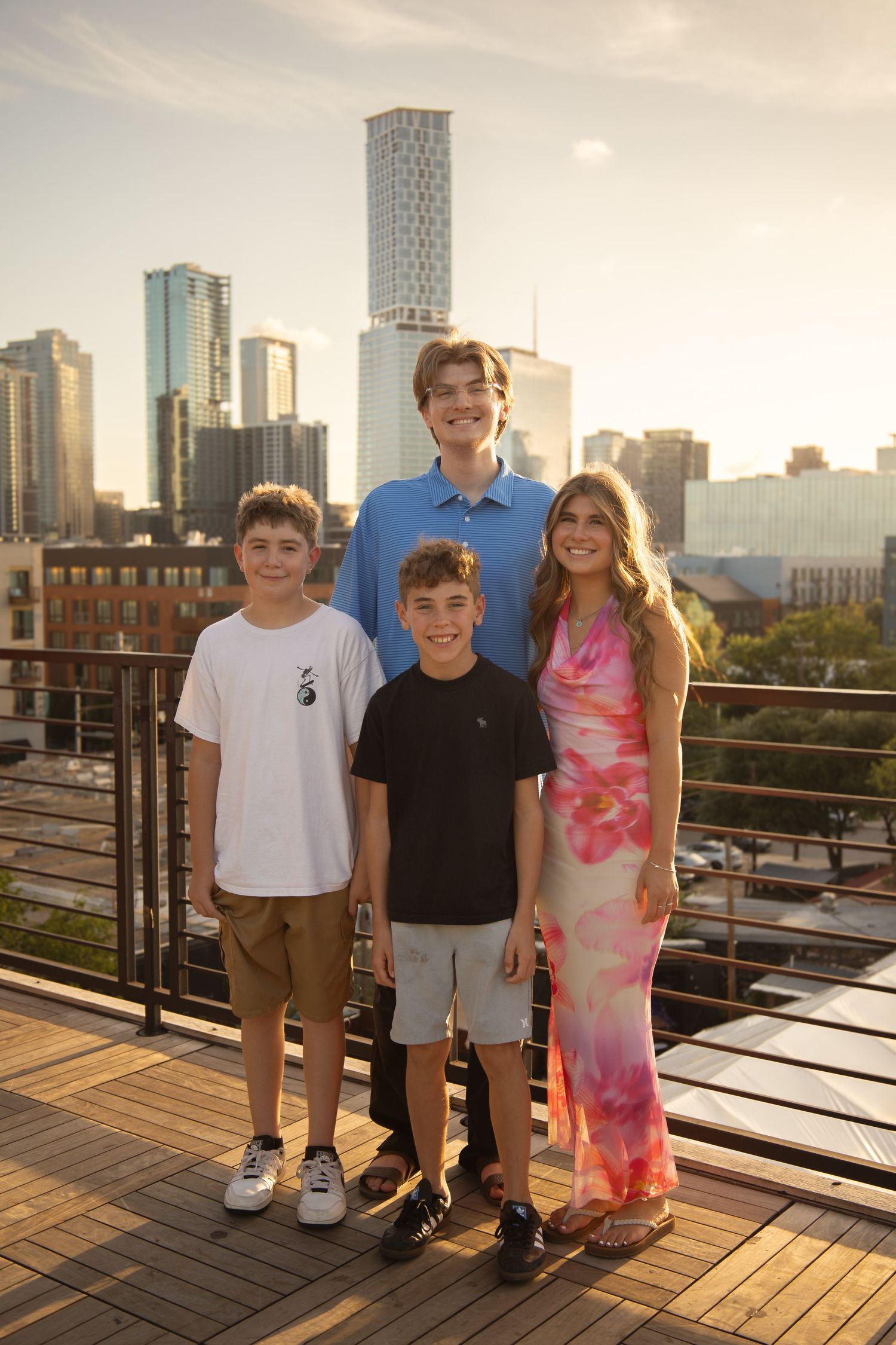 Four young people smiling together on a rooftop deck with a city skyline at golden hour.